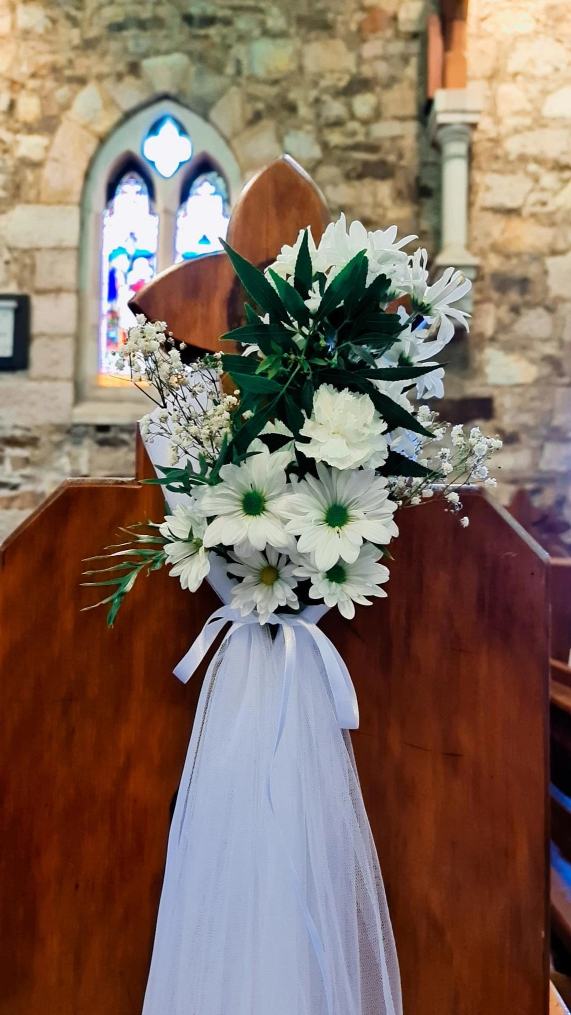 White floral arrangement with tulle bow decorating a wooden church pew in front of a stone wall and stained glass window.