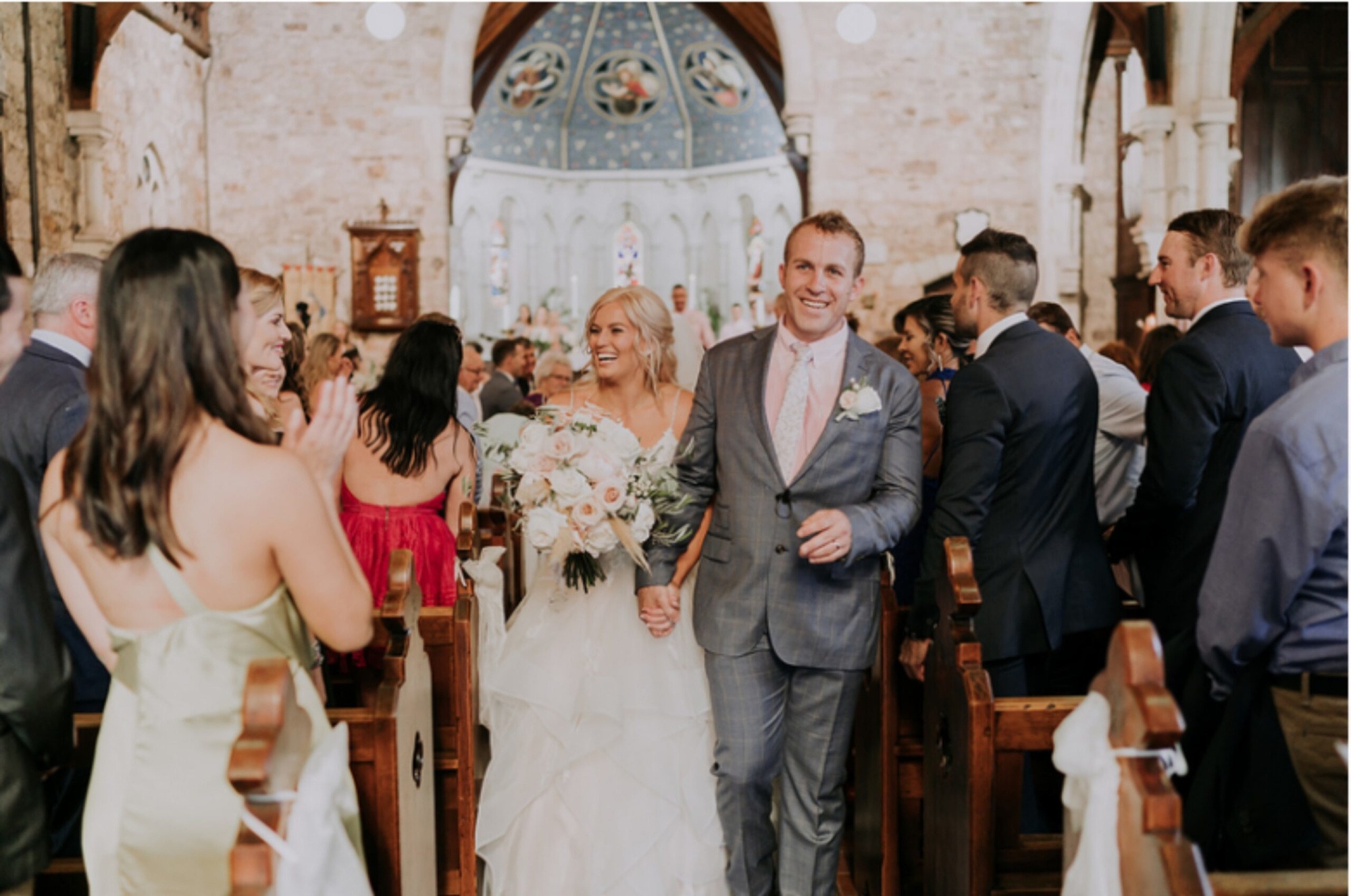 Bride and groom walk back down the aisle smiling as guests applaud inside a stone church.