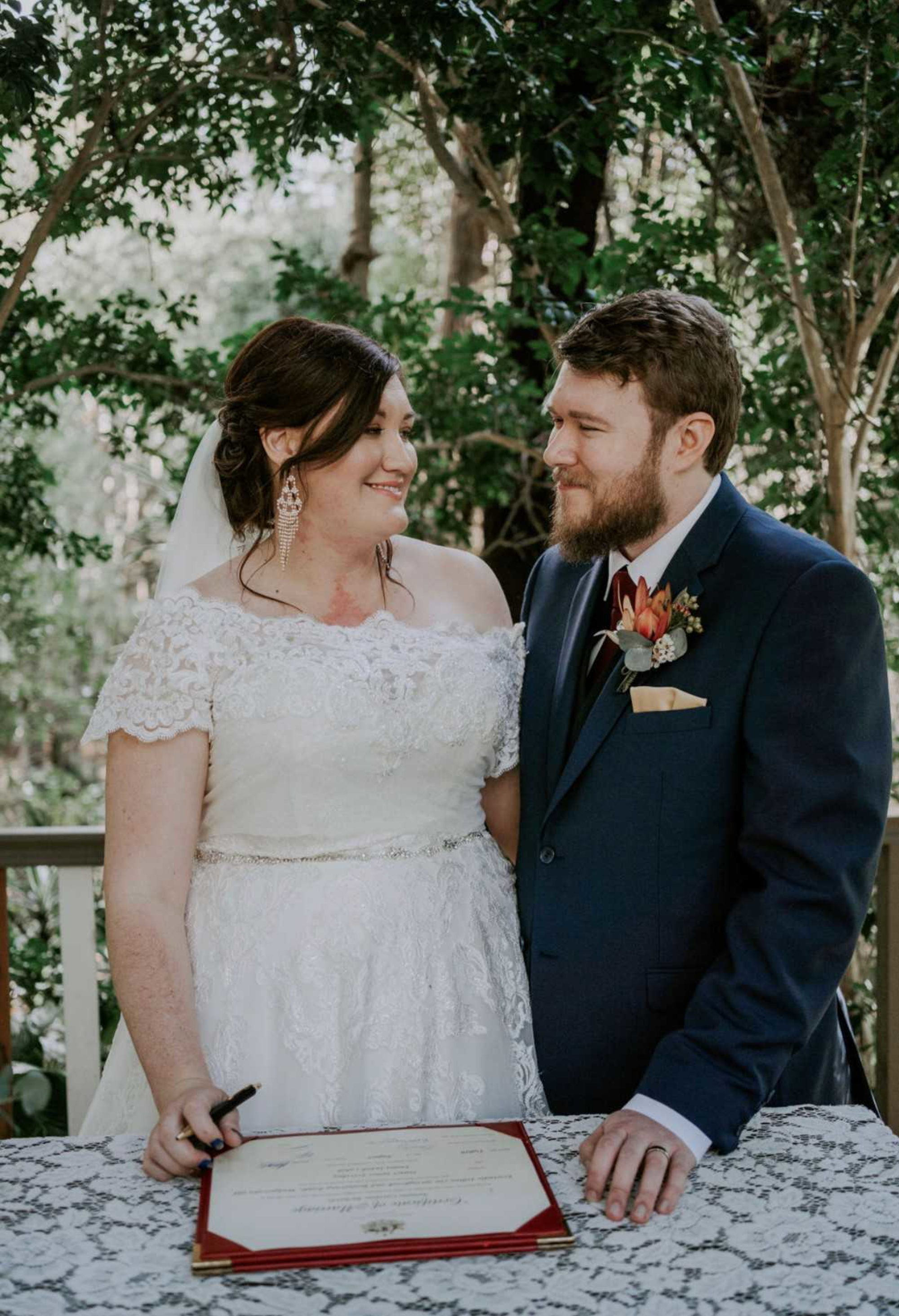 Bride and groom smile at each other while signing their marriage certificate at an outdoor garden wedding.