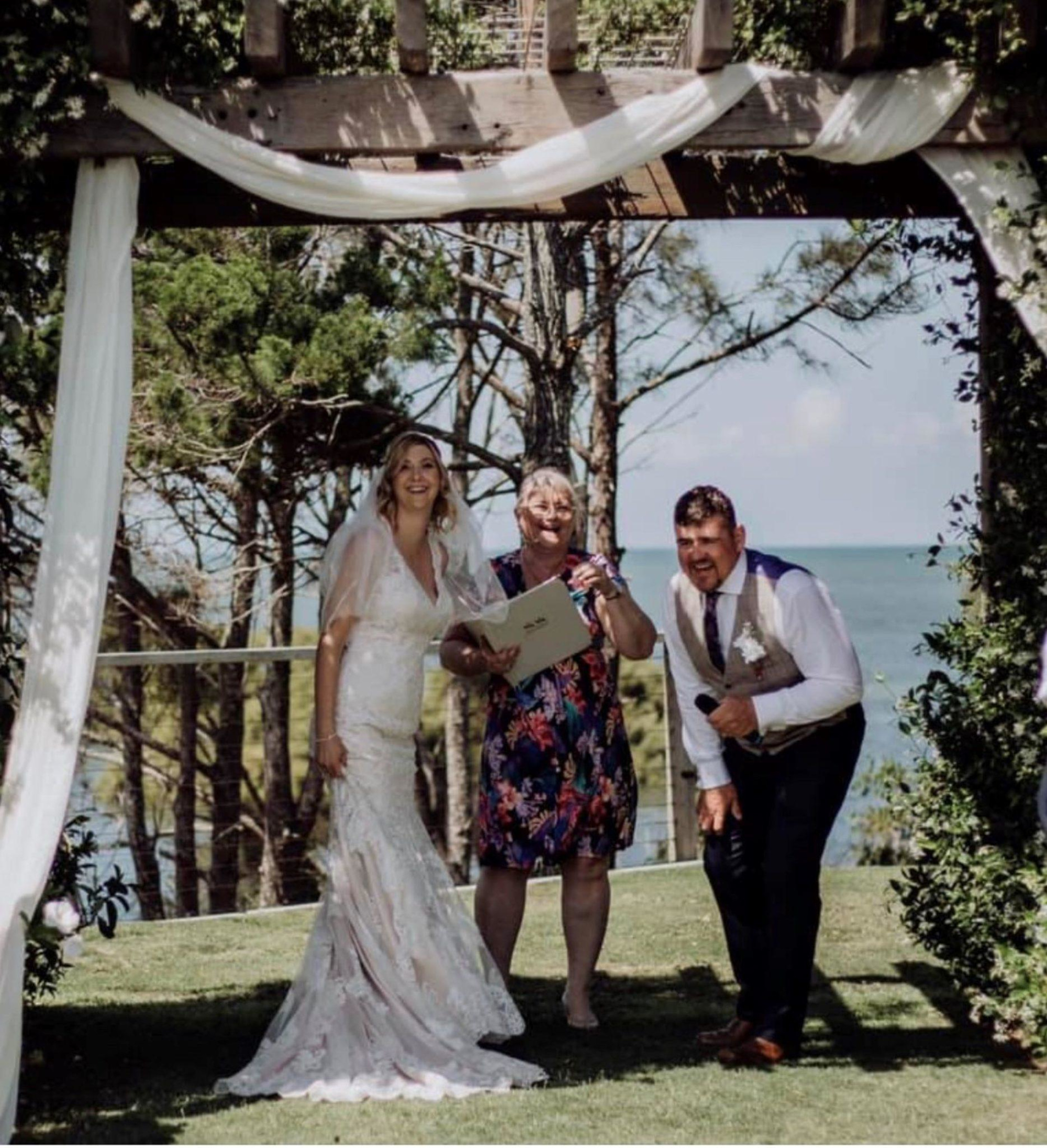 Bride and groom laugh with a celebrant under a draped wooden arch at an outdoor waterfront ceremony.