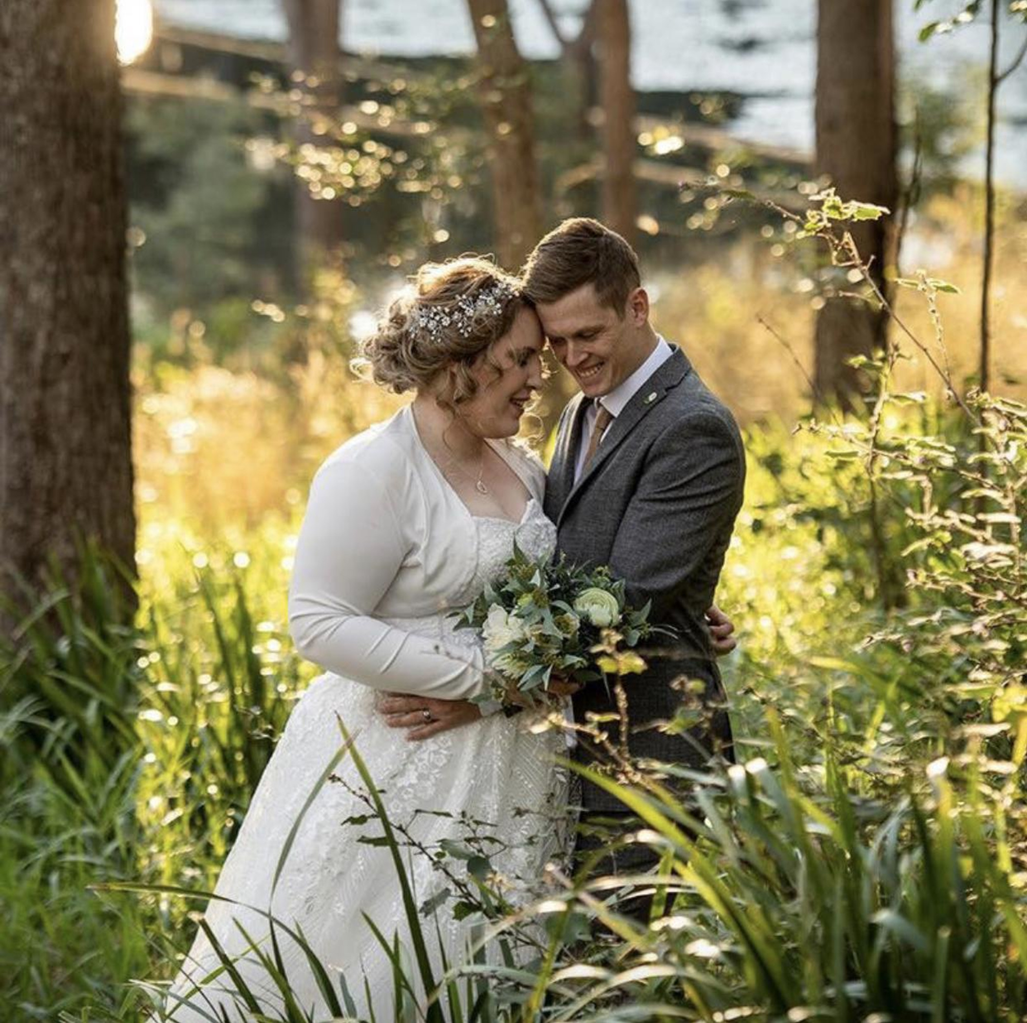 Bride and groom embrace with a bouquet in a sunlit forest clearing.