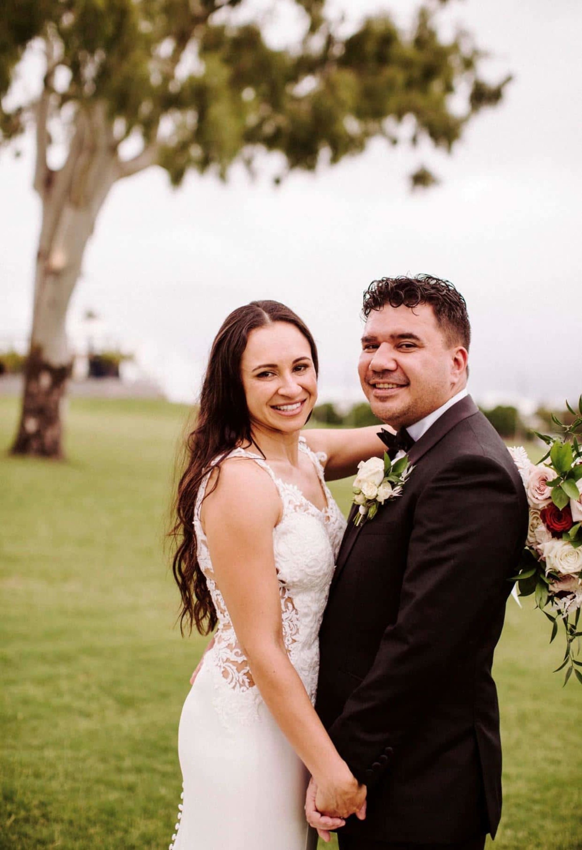 Smiling wedding couple holding hands outdoors with bouquet and greenery in the background.