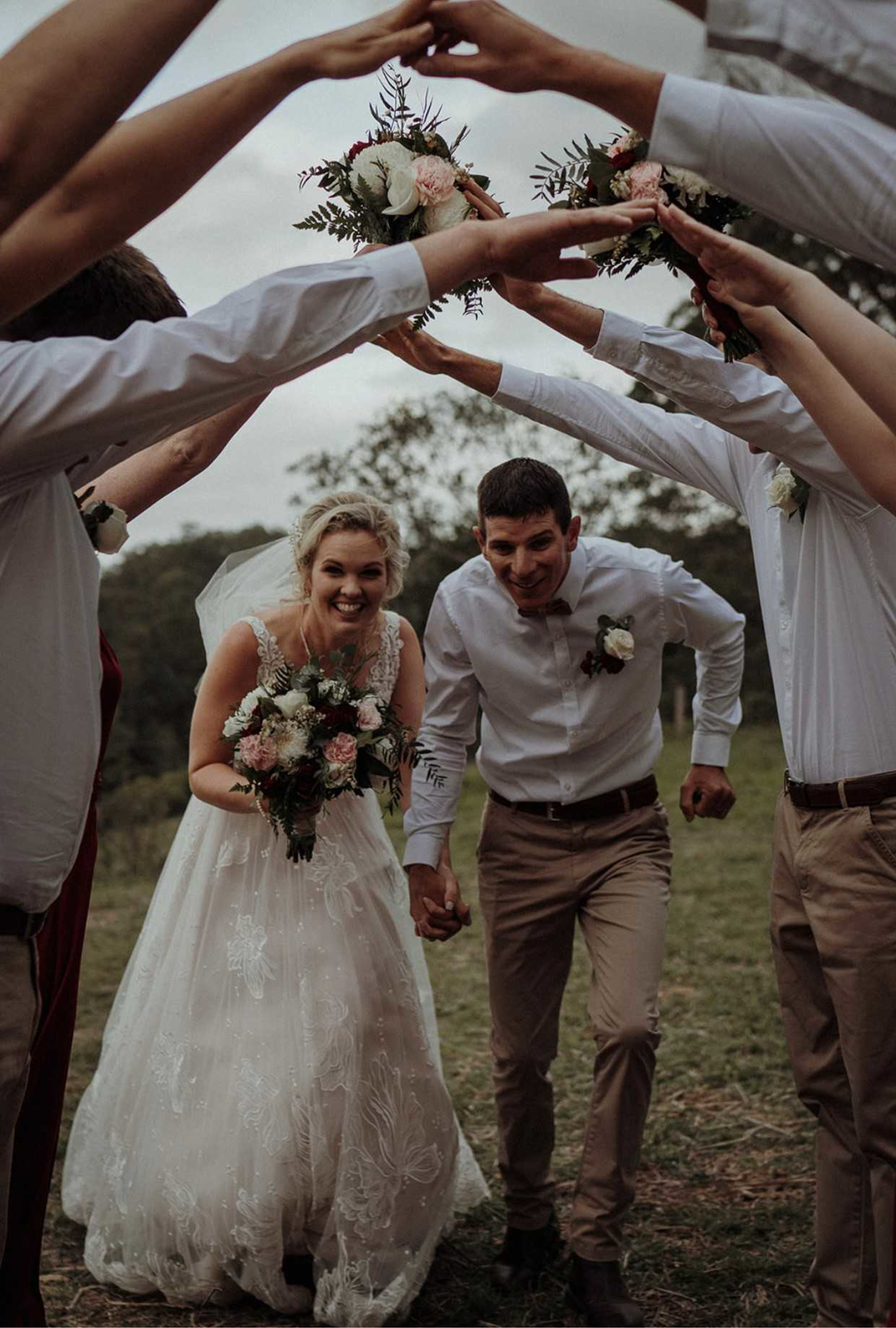 Smiling bride and groom run hand in hand under an arch of raised arms and bouquets at an outdoor wedding.