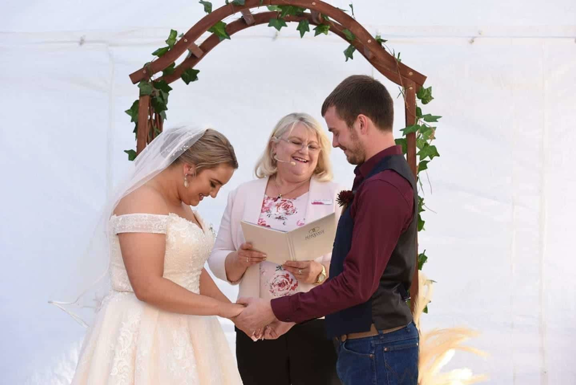 A wedding celebrant leads a joyful outdoor ceremony as the bride and groom hold hands beneath a rustic arch.