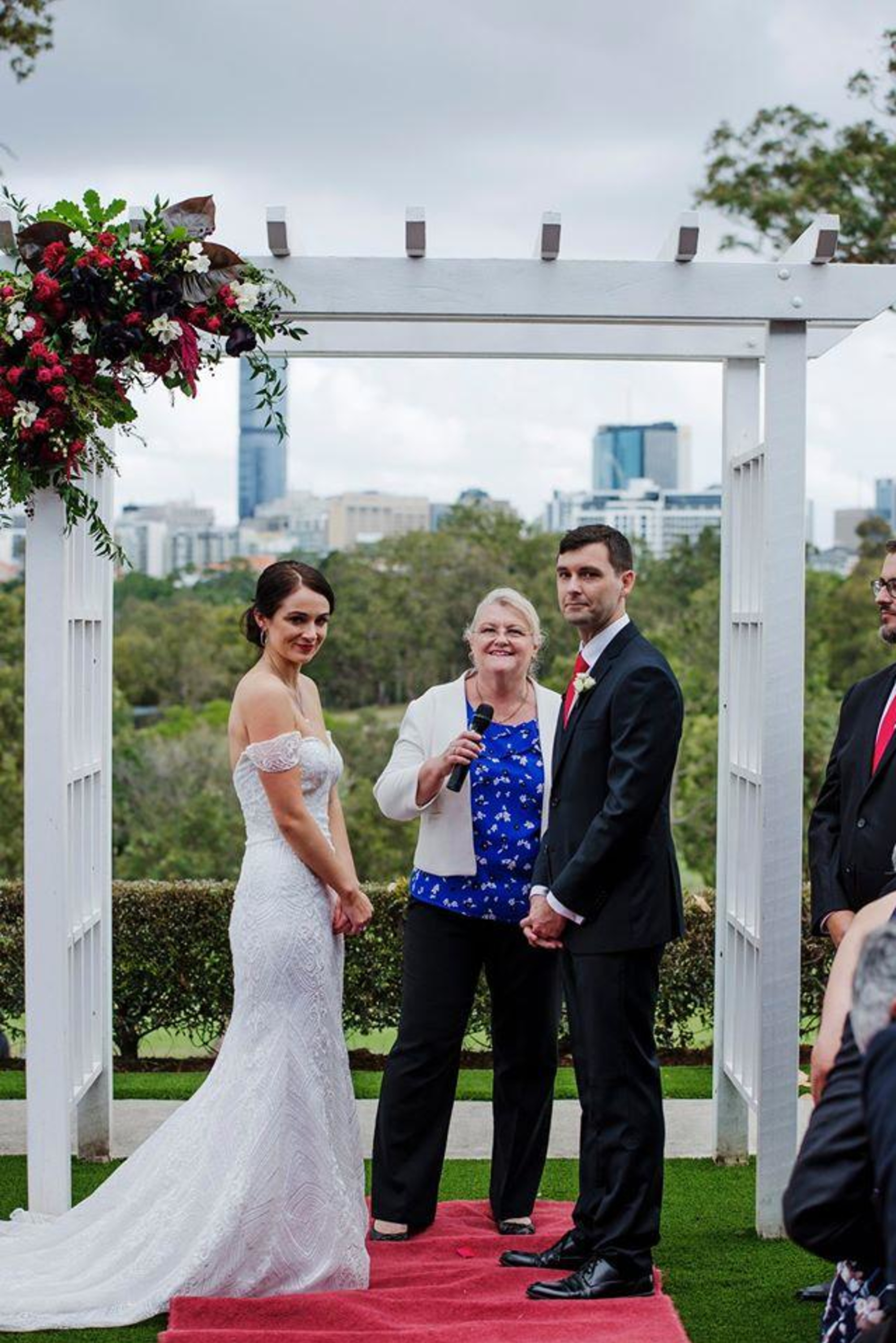 Bride and groom stand with their celebrant under a floral wedding arbor overlooking a city skyline.