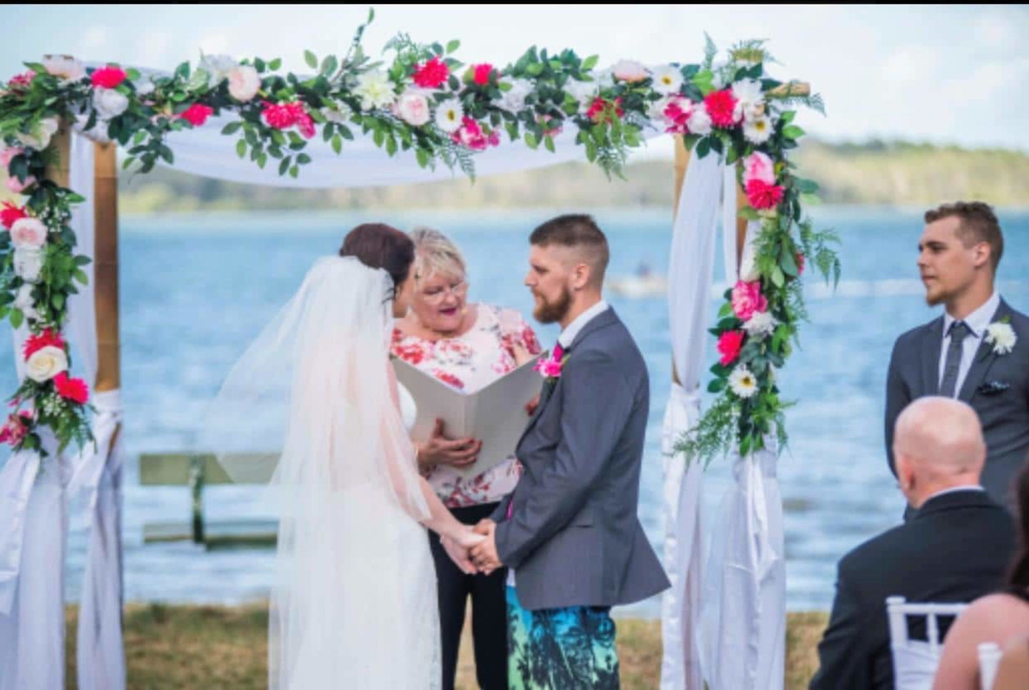 Couple holding hands under a floral wedding arch during an outdoor waterfront ceremony.