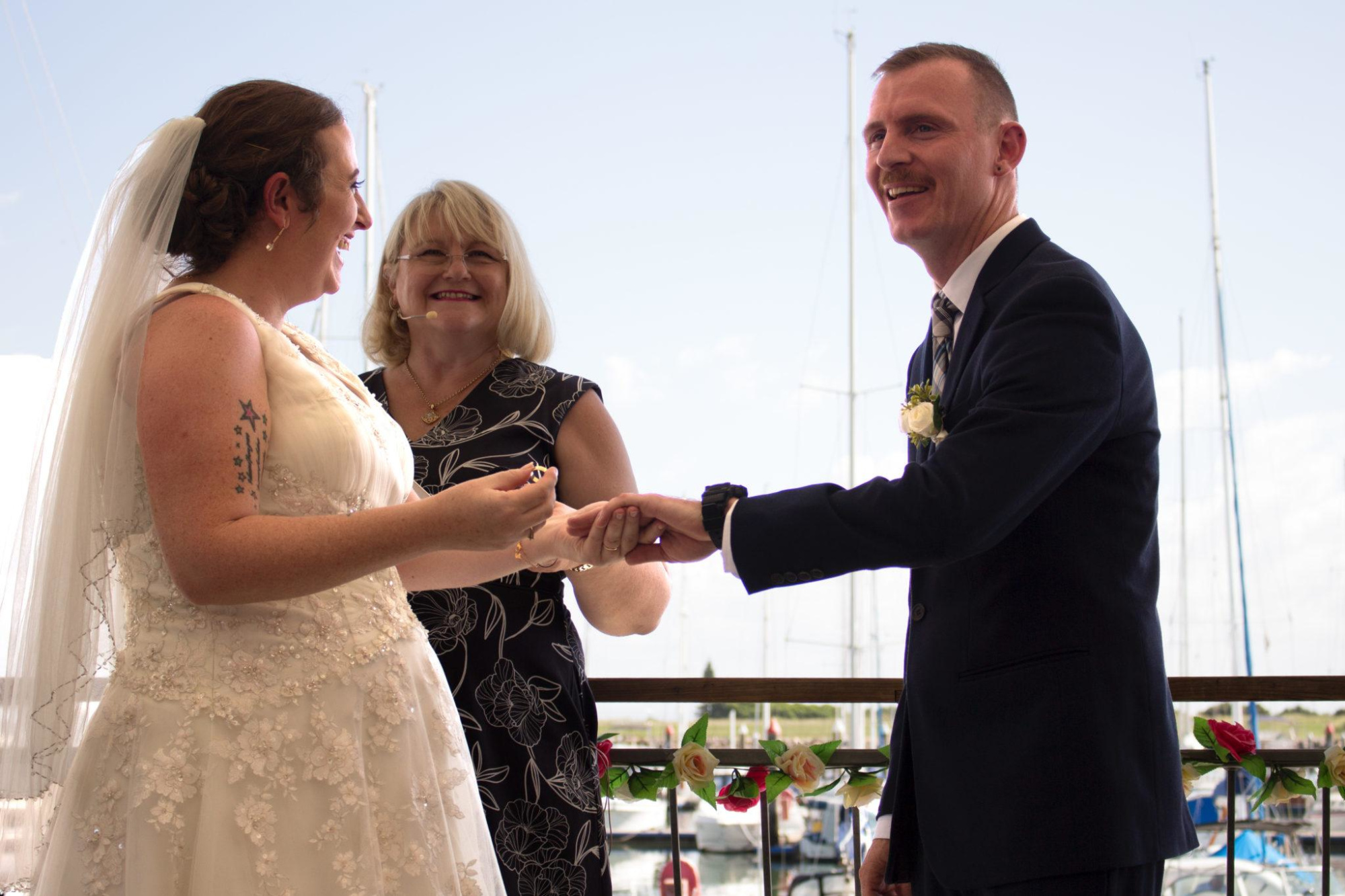 Bride and groom exchange rings during an outdoor marina wedding ceremony with a celebrant smiling beside them.