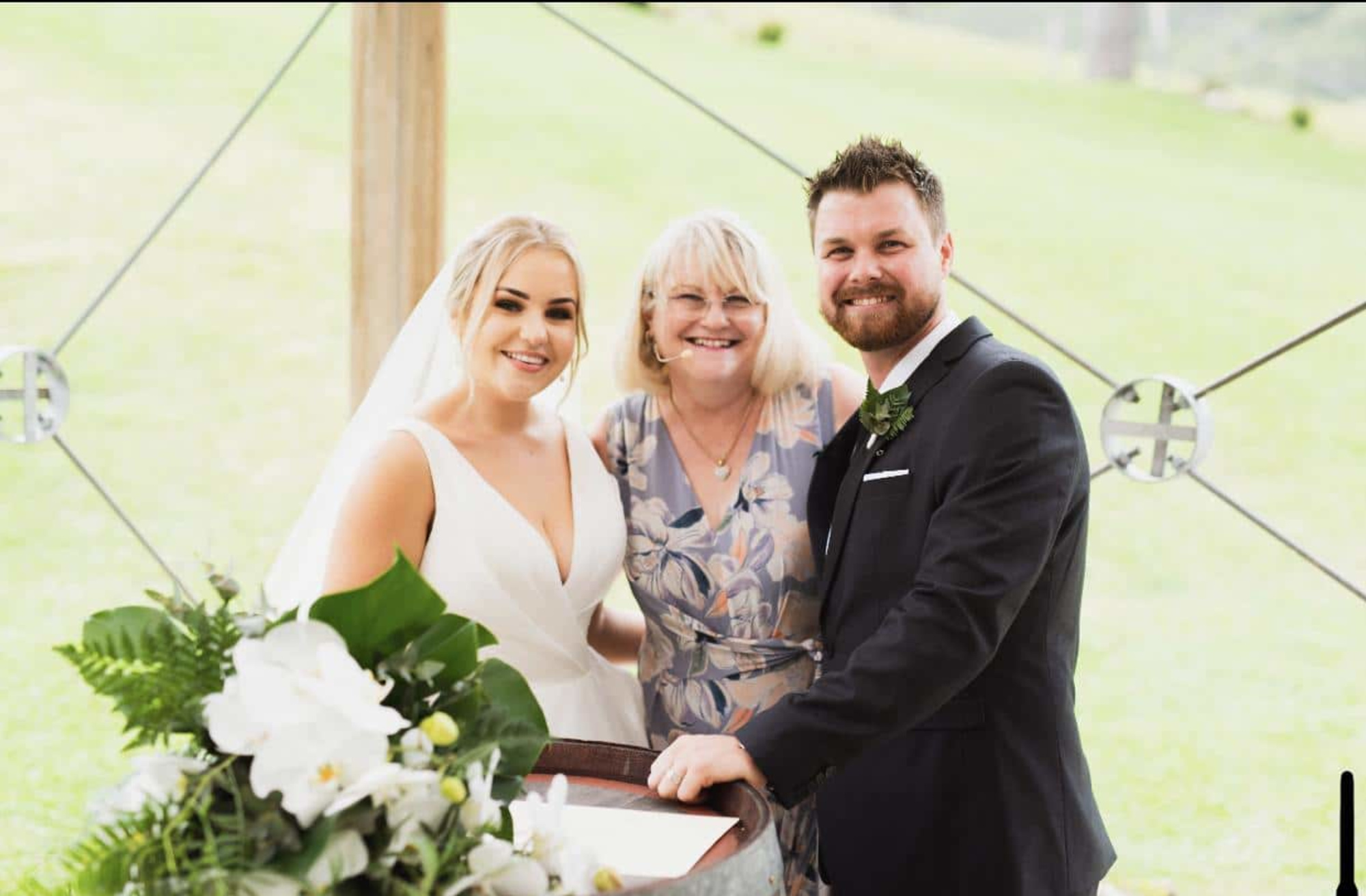 Bride and groom smiling with their celebrant during an outdoor wedding ceremony beside a floral arrangement.