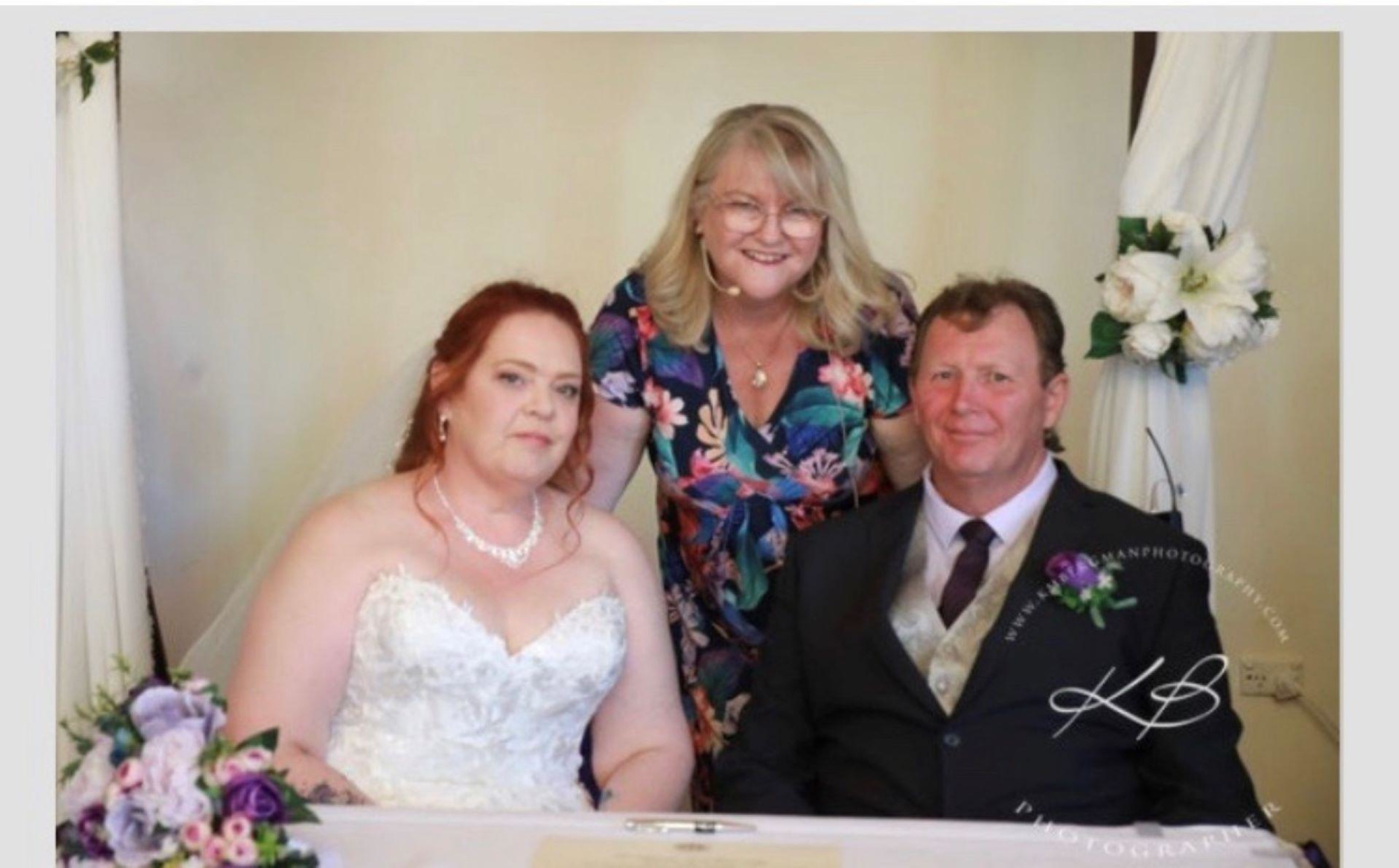 Bride and groom sit at the signing table with their celebrant standing behind them, framed by floral decor.
