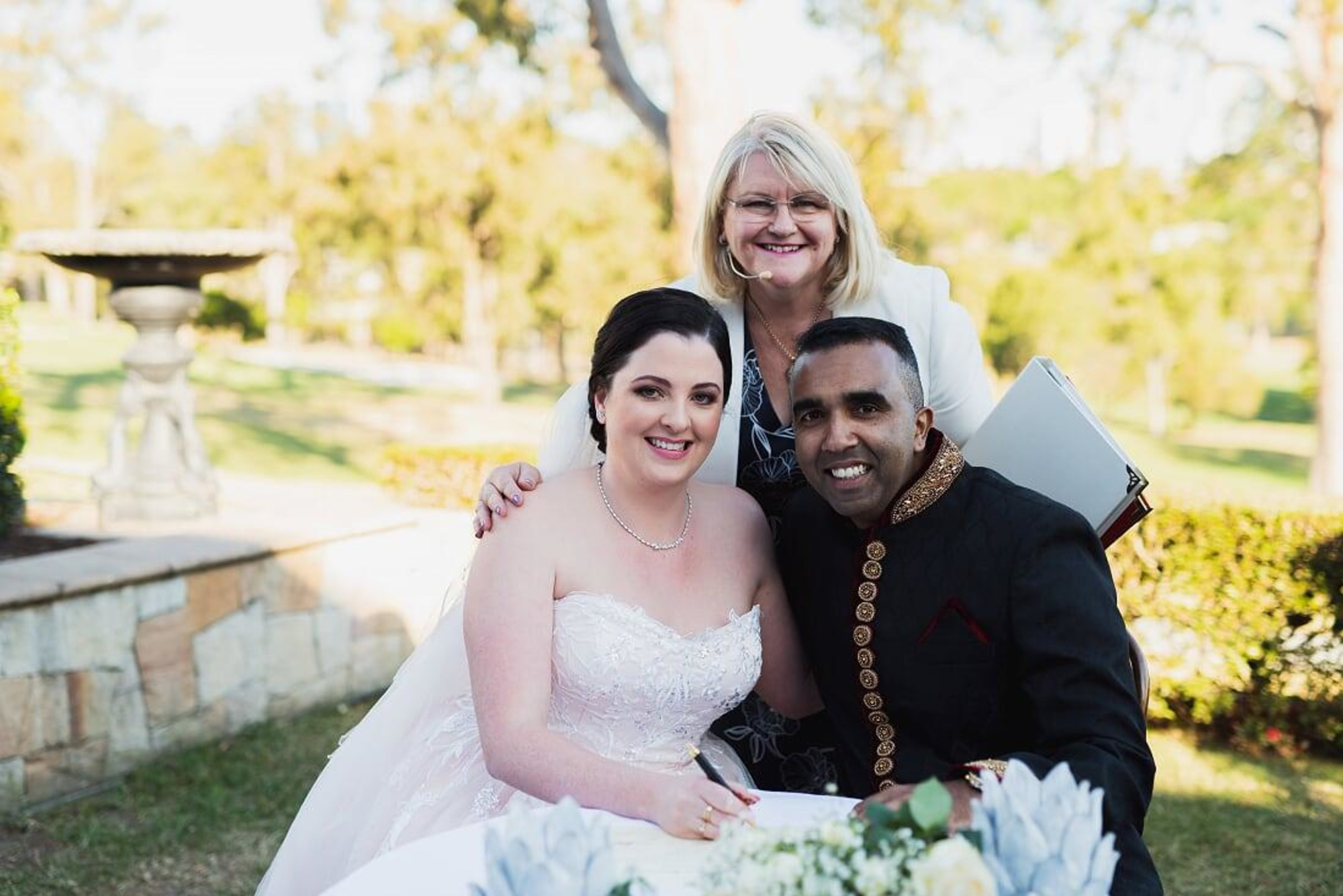 Bride and groom smiling with their celebrant while signing the marriage register at an outdoor garden wedding.