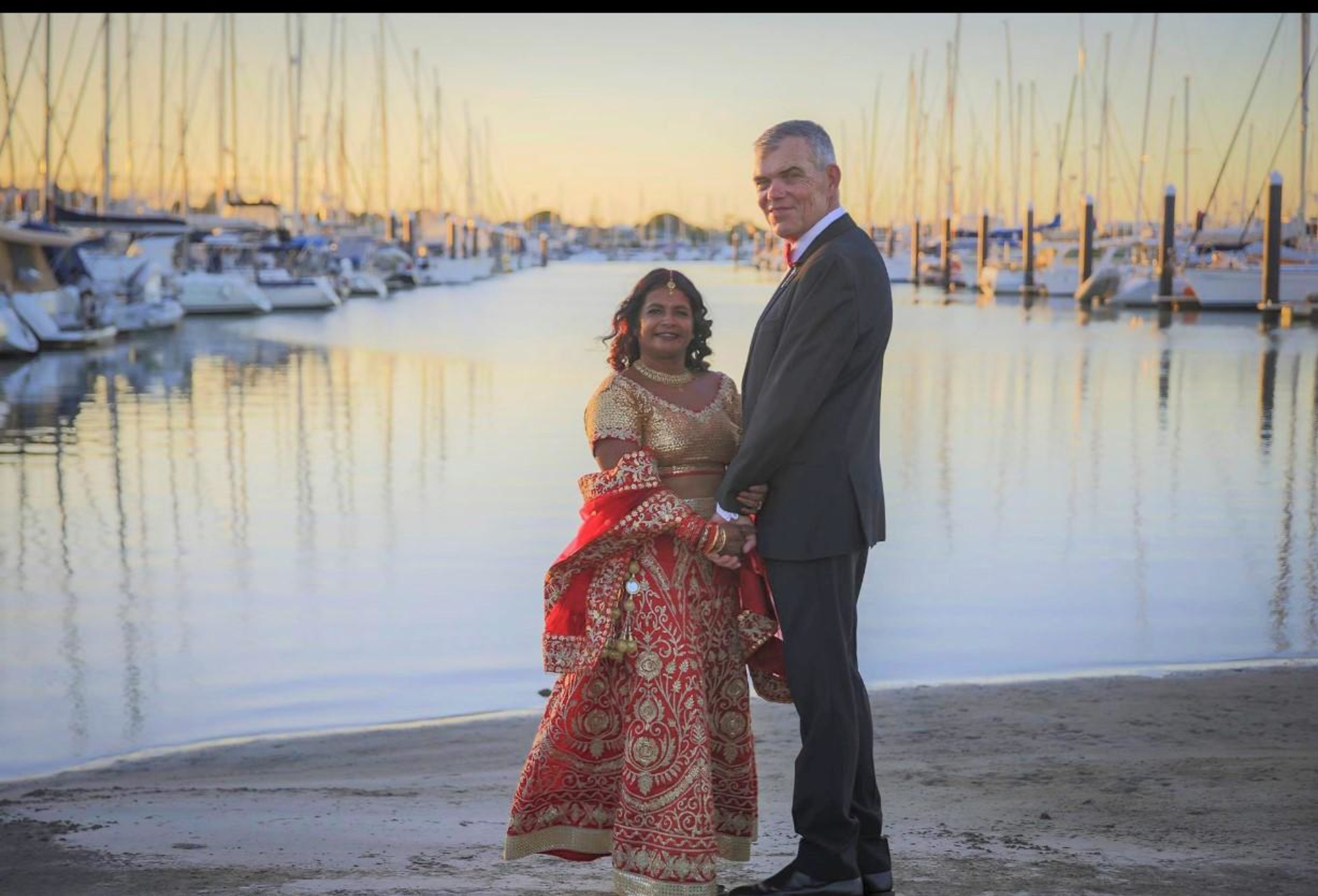 Wedding couple posing on a sandy marina shoreline at sunset with boats and calm water in the background.