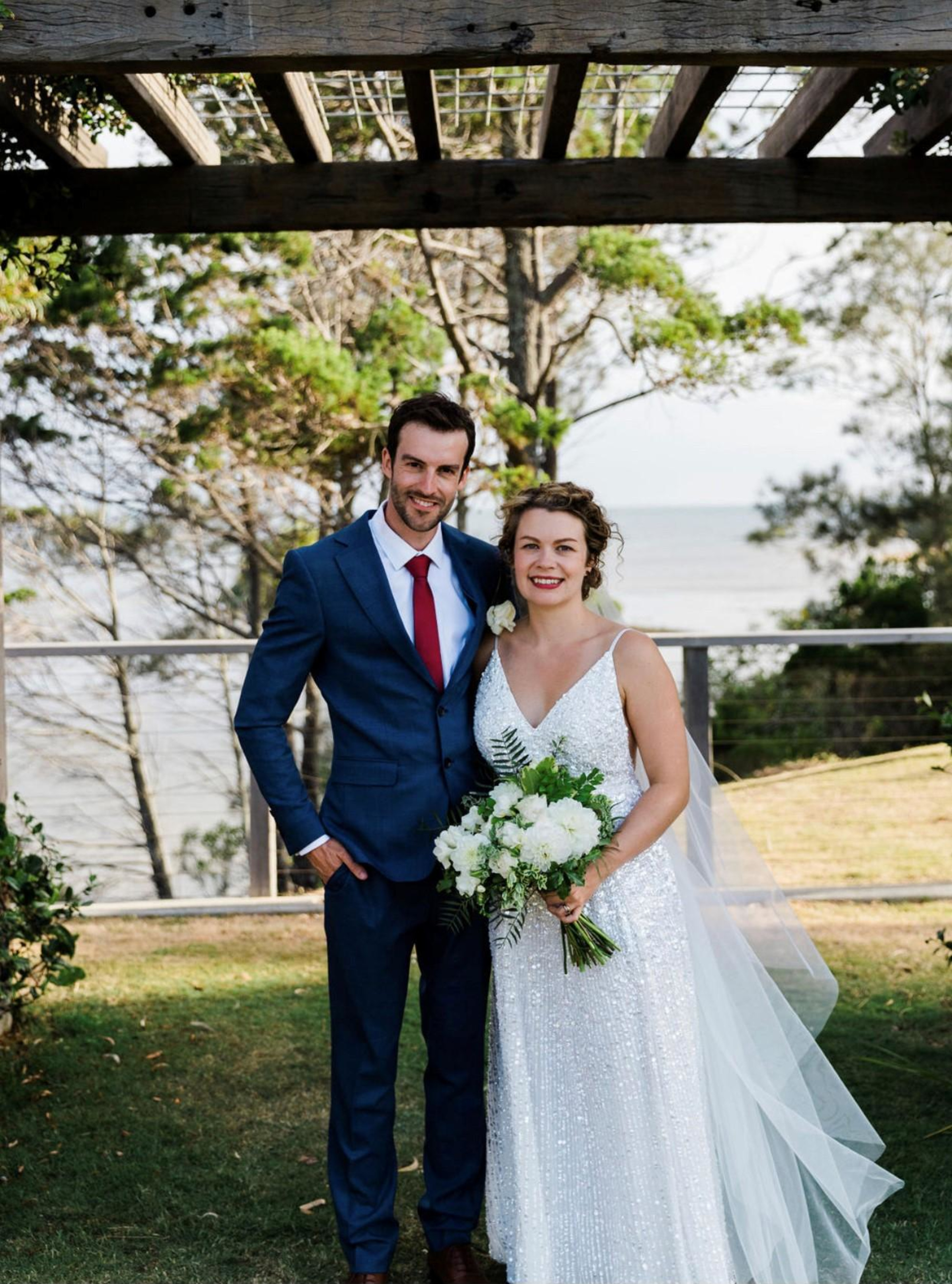 Bride and groom posing under a wooden arbor at an outdoor waterfront wedding.