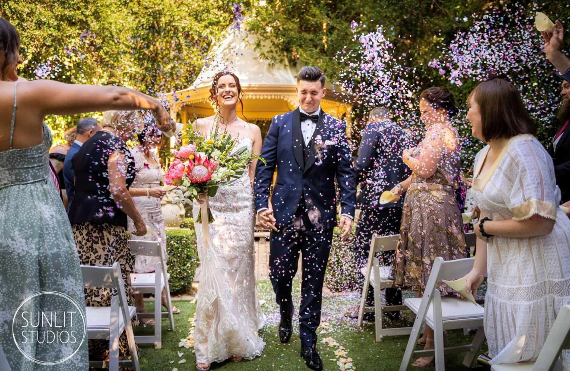 Newlywed couple walks down the aisle in a garden as guests shower them with colorful confetti.