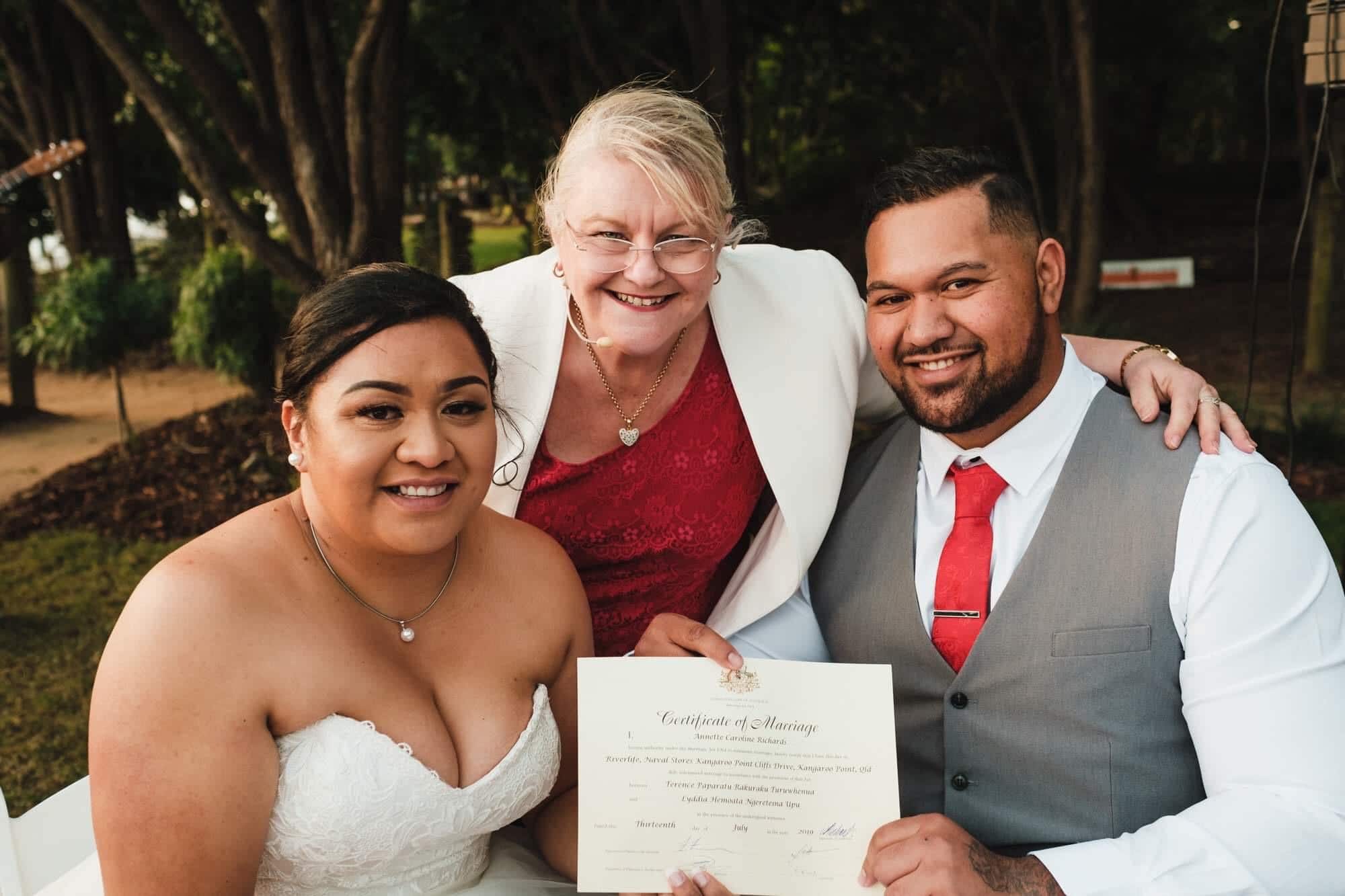 Smiling wedding couple with their celebrant holding a signed marriage certificate outdoors.