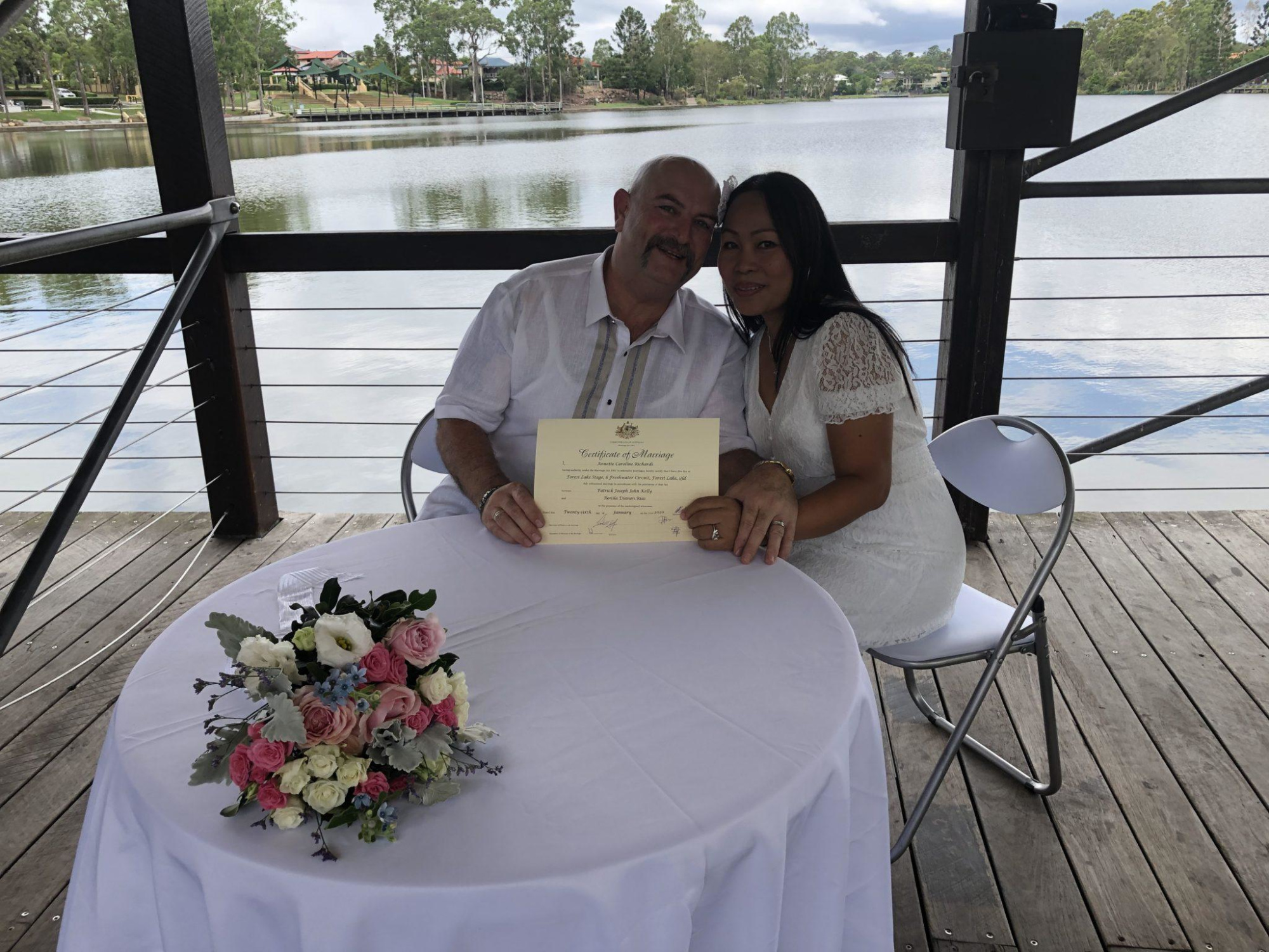Couple in white outfits sit by a lakeside table holding their marriage certificate with a pastel bouquet in front of them.