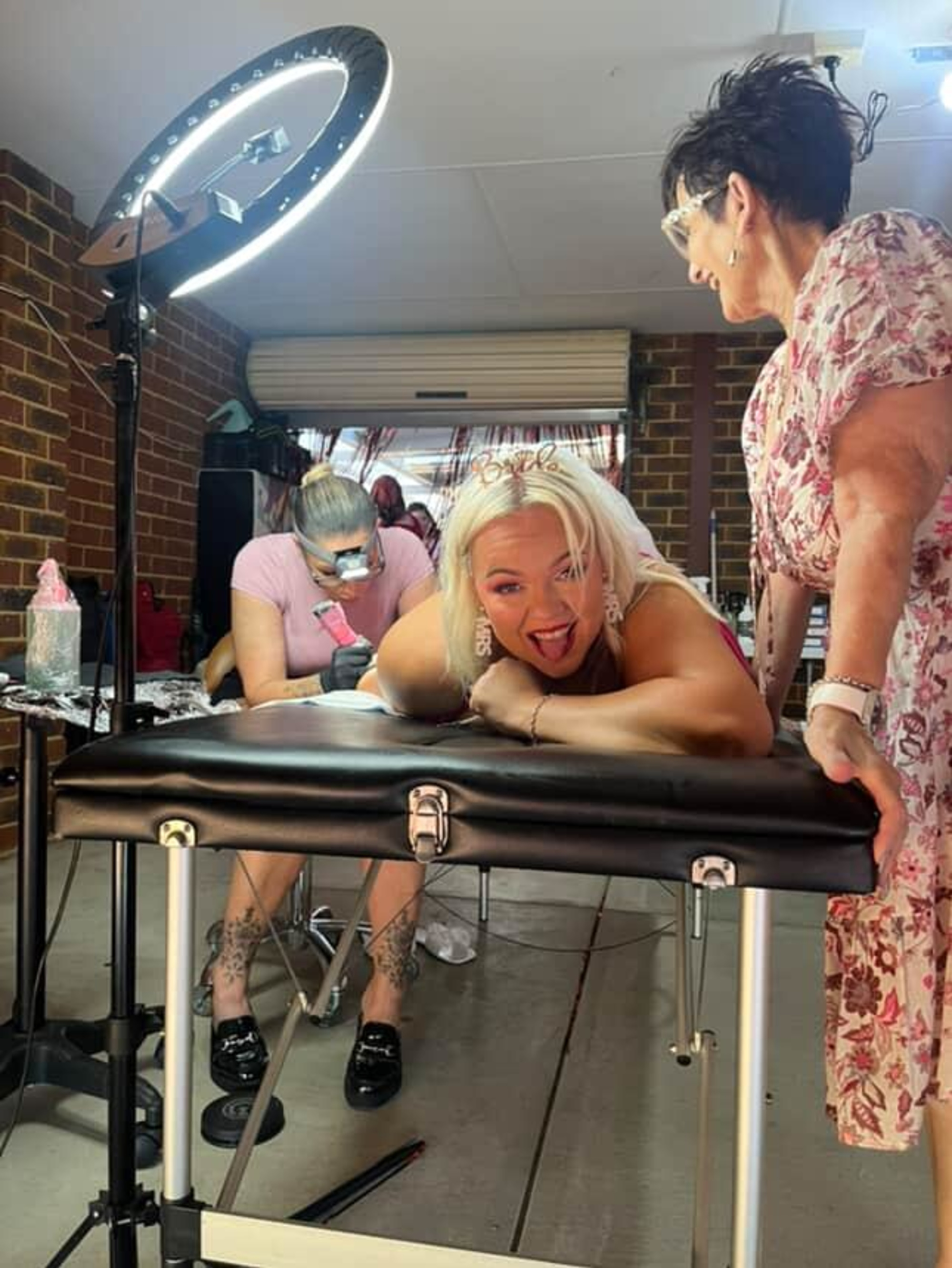 Bride wearing a tiara gets a tattoo while laughing with a friend in a studio setup.