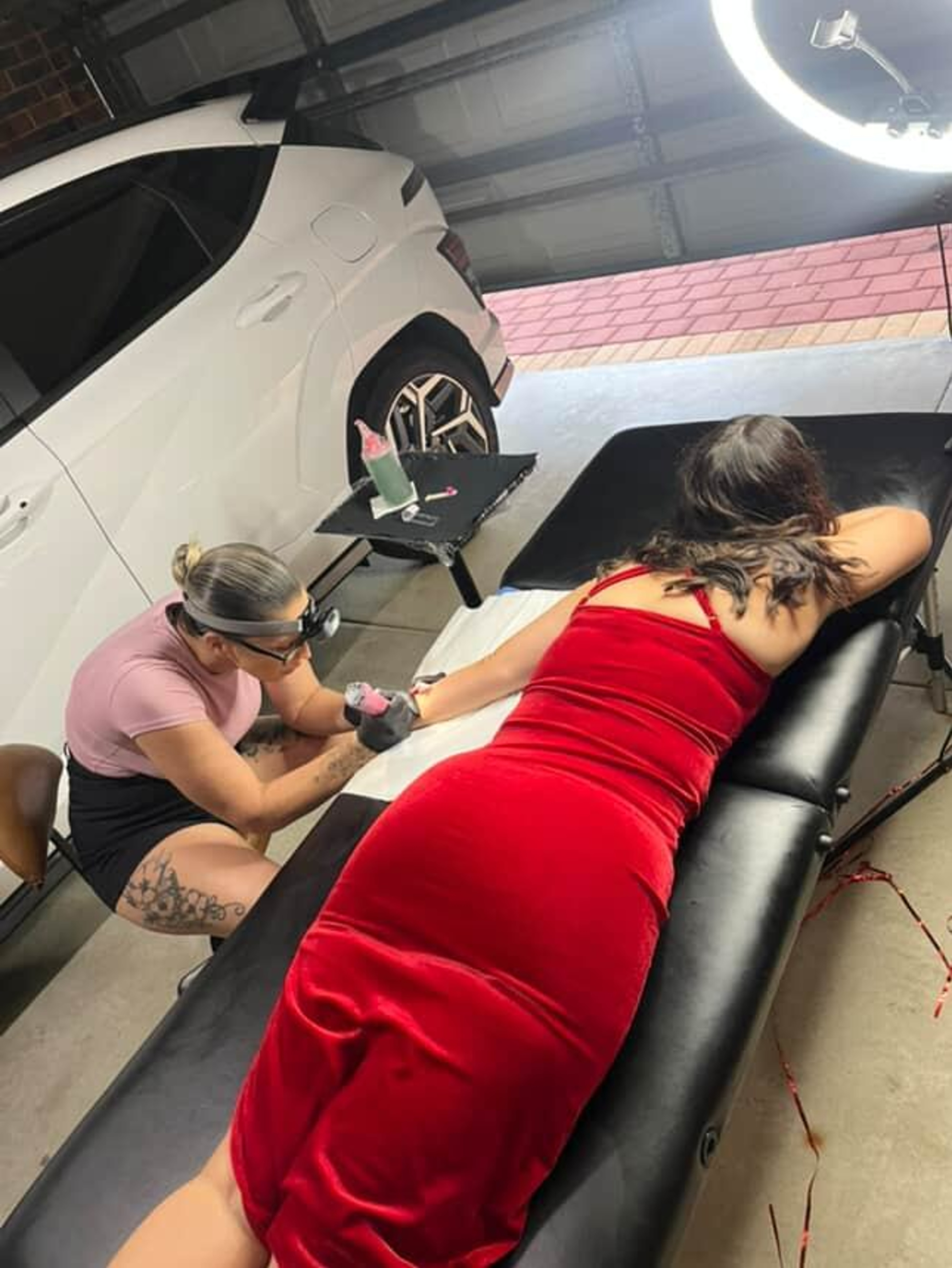 Tattoo artist working on a woman in a red dress lying on a studio table beside a white car.