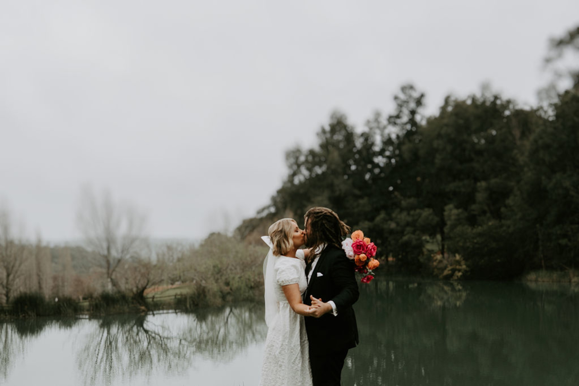 Wedding couple kisses by a tranquil lakeside with a bright bouquet against a forest backdrop.