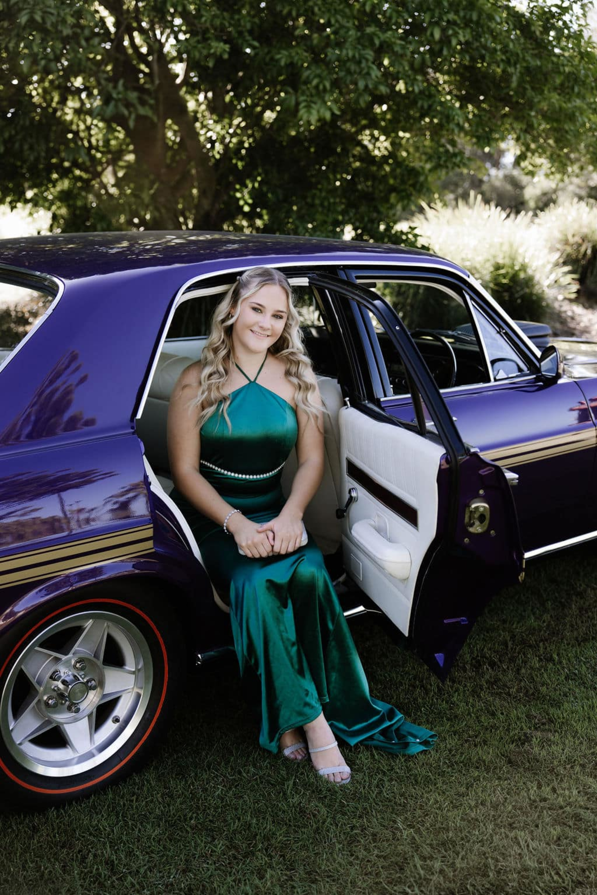 Woman in an emerald gown sits in the open door of a purple classic car on a grassy outdoor setting.