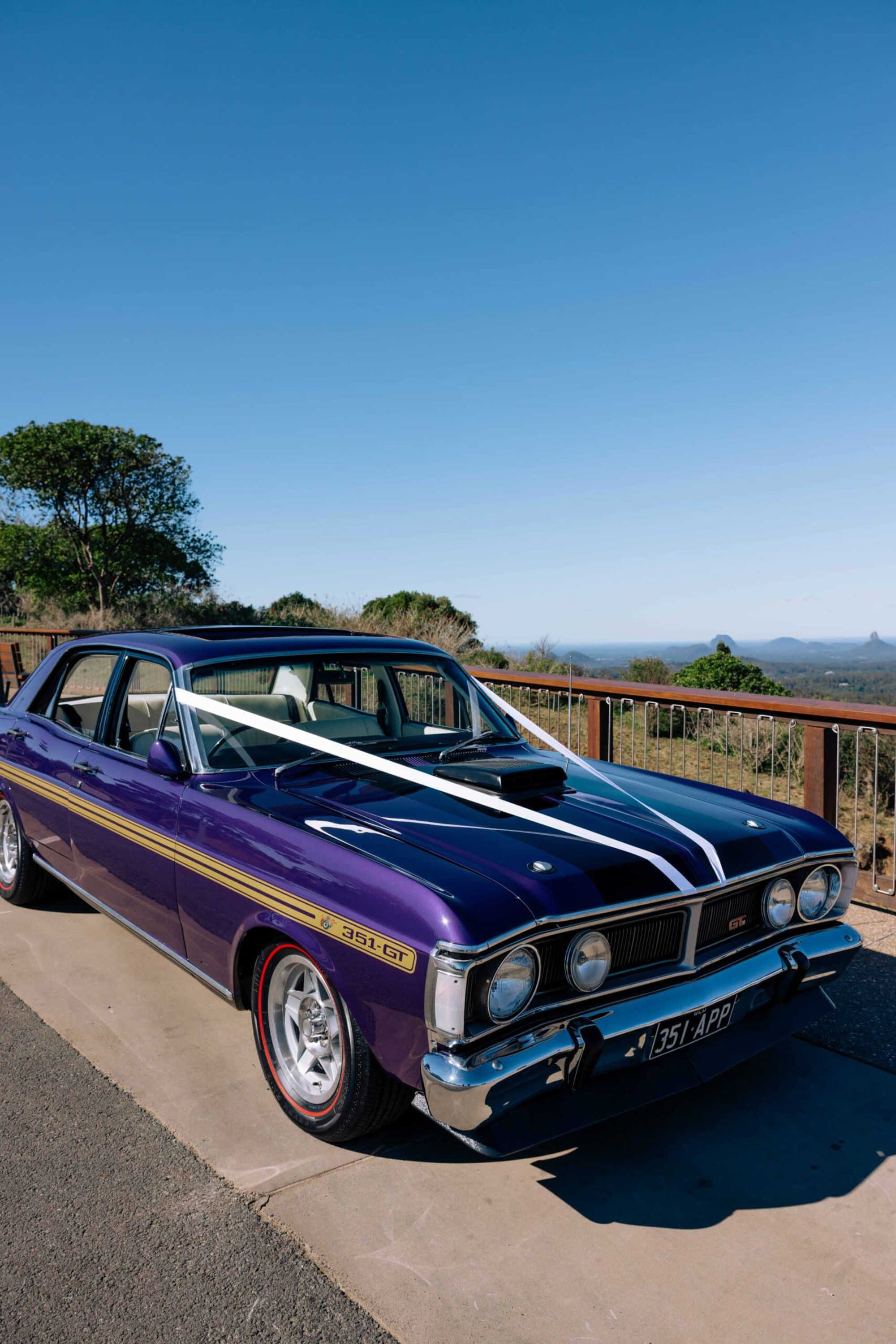 Purple vintage wedding car with white ribbons parked at a scenic lookout under a clear blue sky.