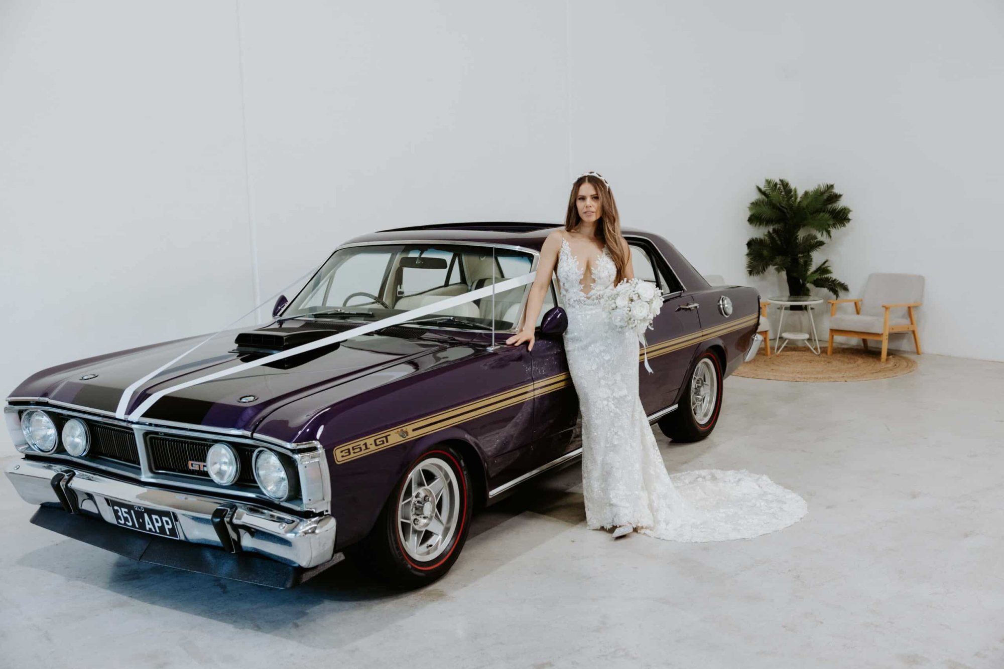 Bride in a lace gown posing with a decorated classic purple wedding car in a minimalist indoor space.