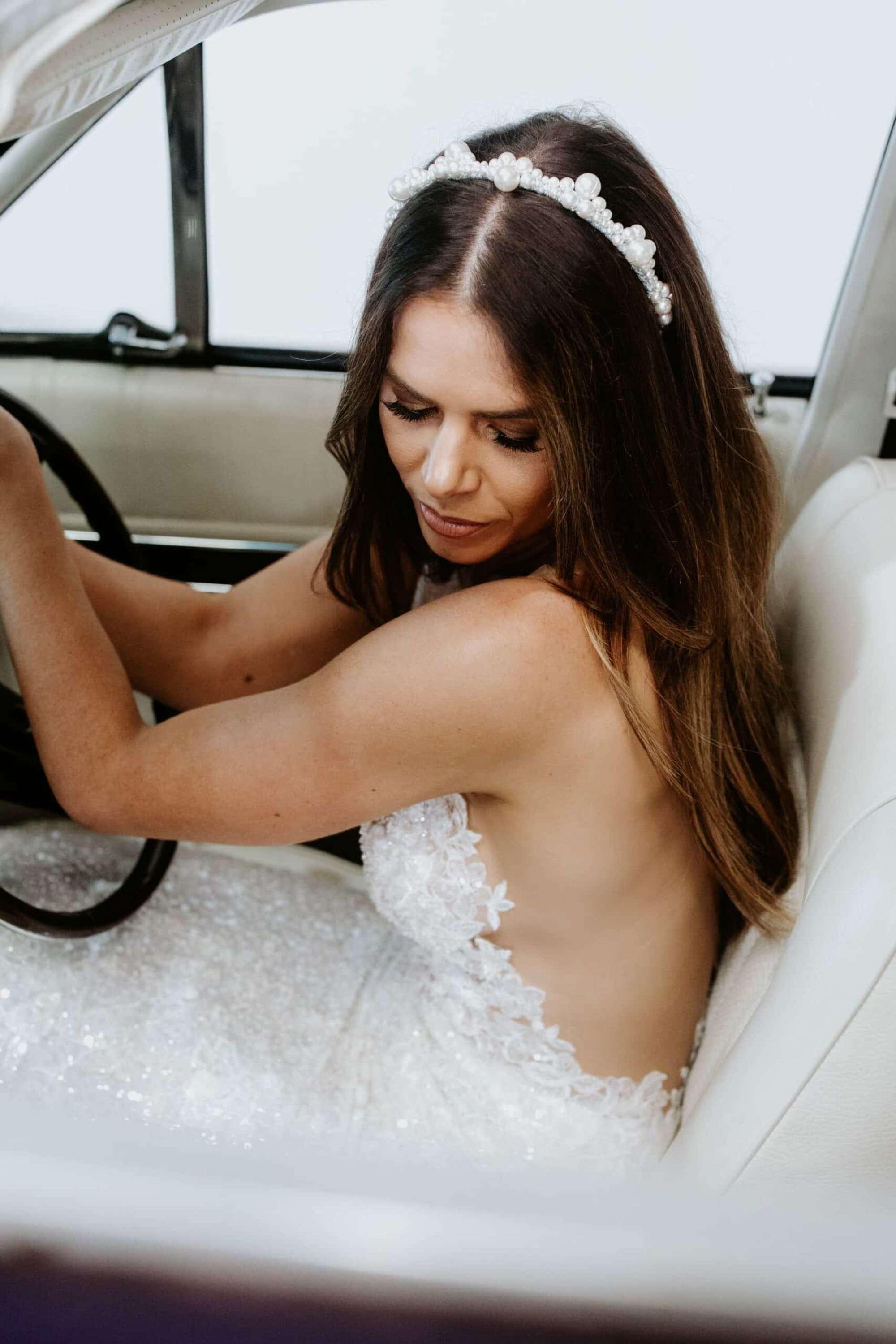 Bride in a beaded gown and pearl headband sitting in a classic car looking down.