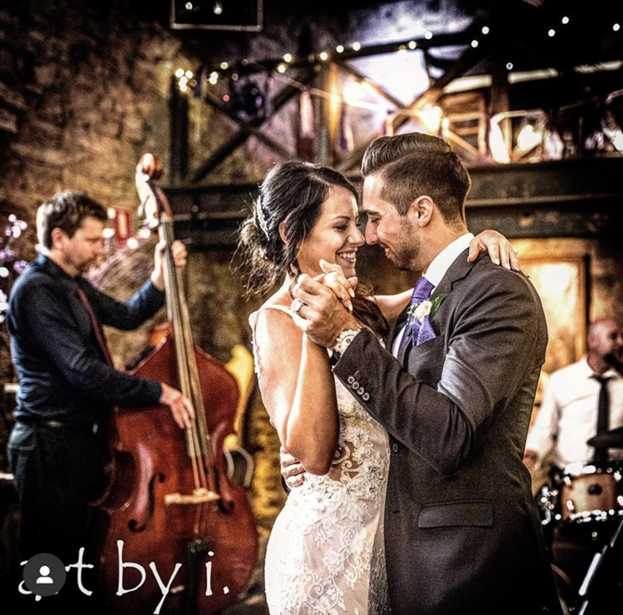 Bride and groom share their first dance at a rustic indoor reception with a live band playing in the background.