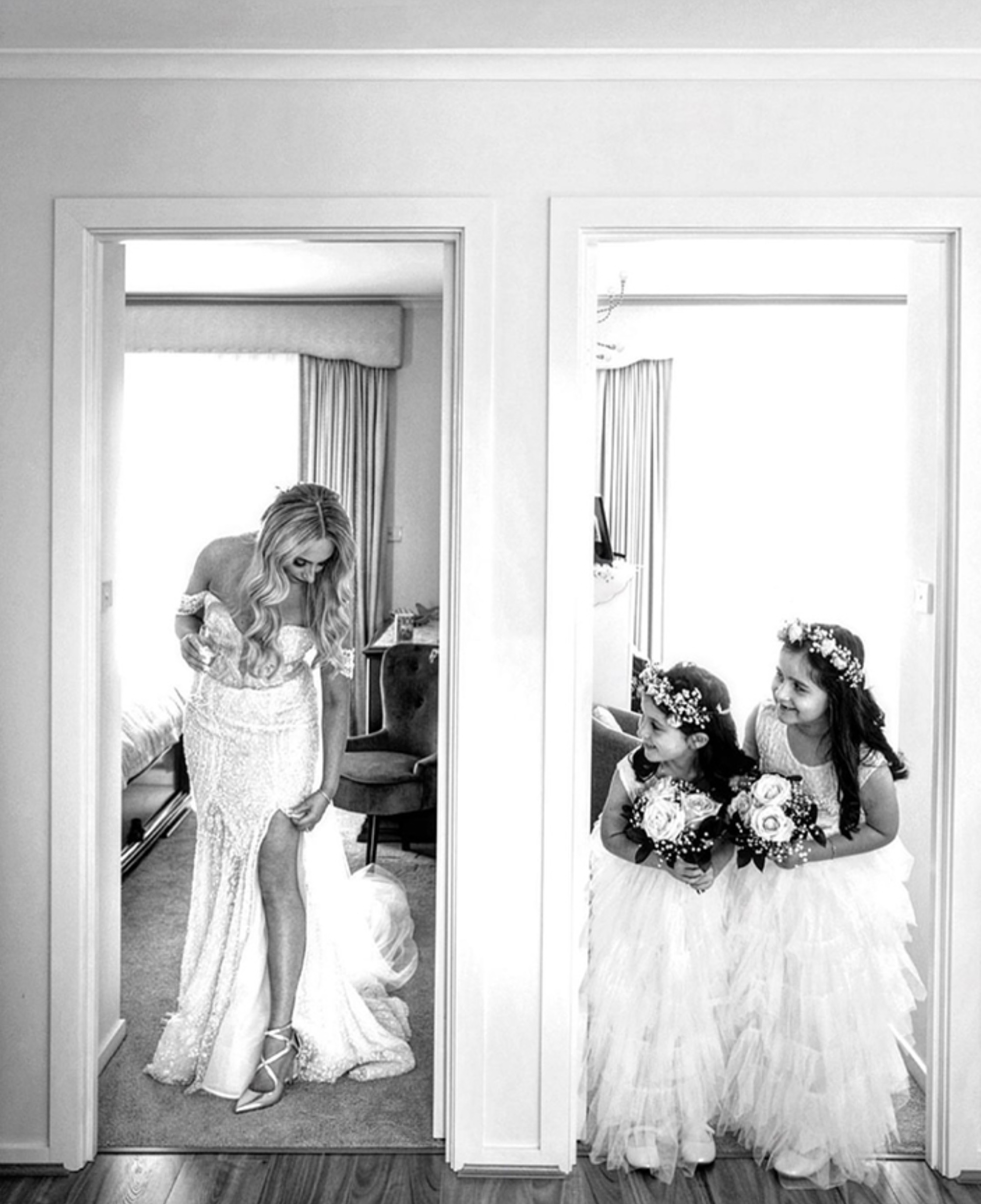 Black and white photo of a bride adjusting her dress as two smiling flower girls watch from the doorway.