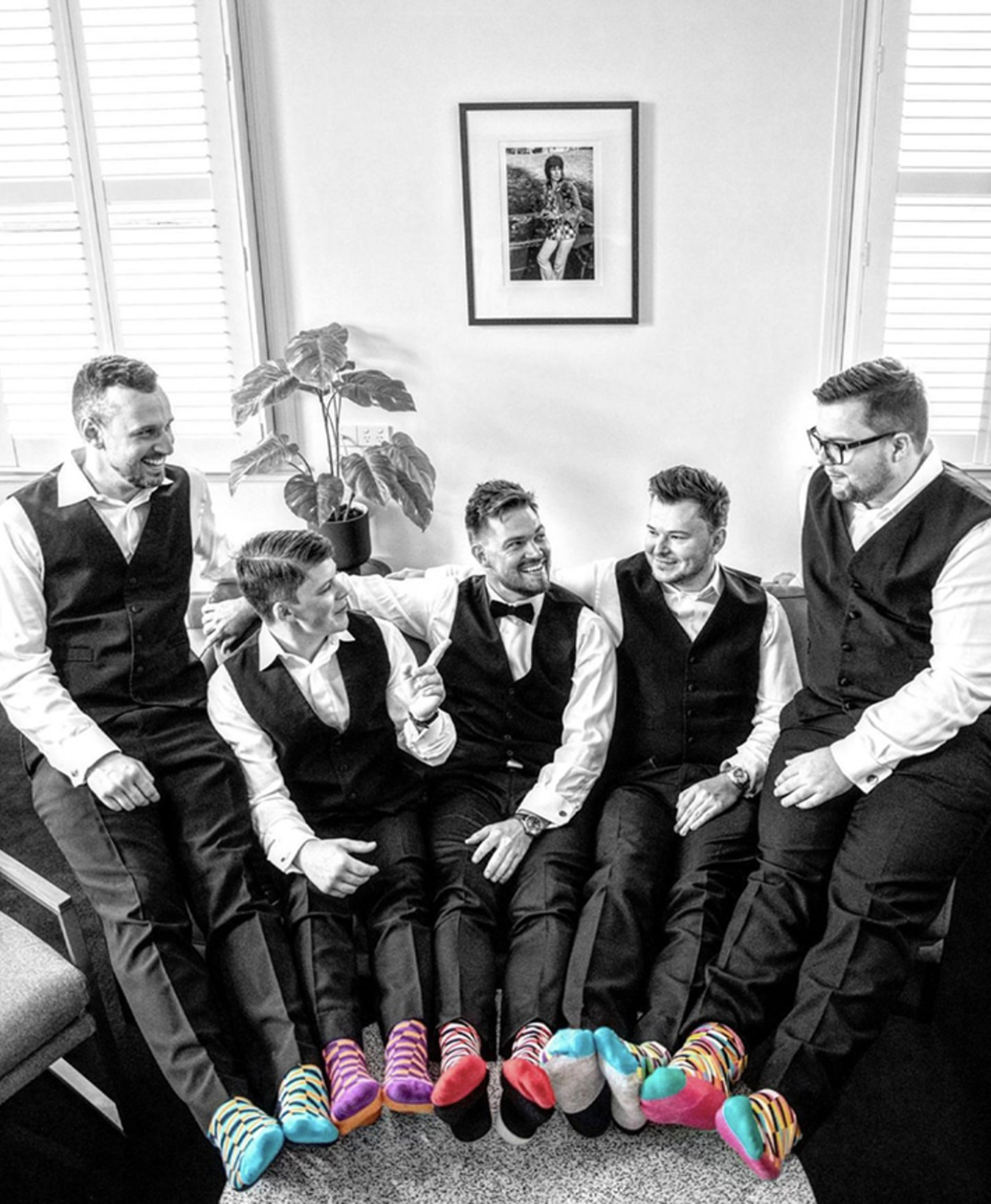 Groomsmen in vests laugh together on a couch, showing off colorful striped socks in an otherwise black and white room.
