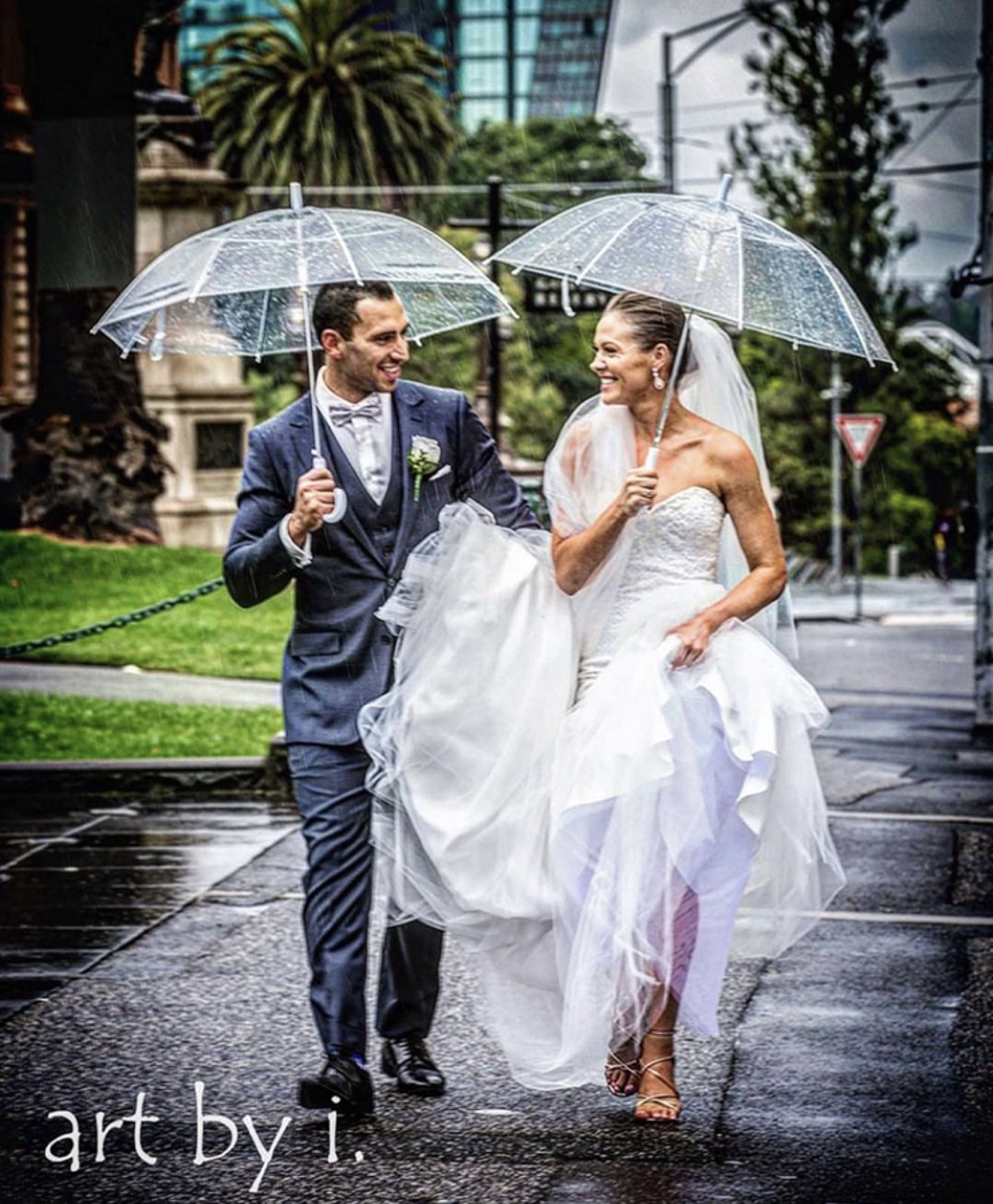 Bride and groom walk under clear umbrellas on a rainy city street, smiling at each other in wedding attire.