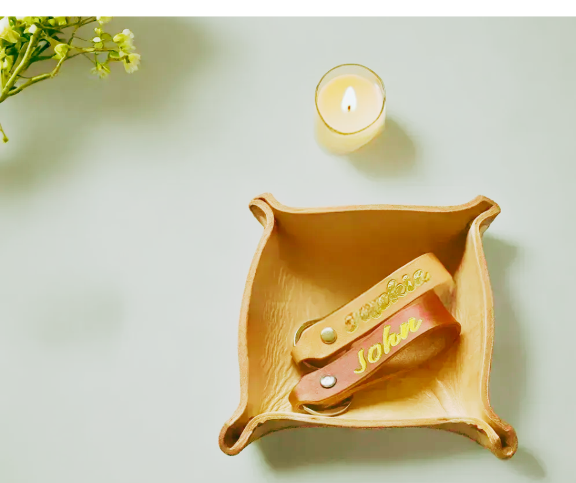 Personalized leather keychains in a small tray beside a candle and greenery on a light surface.
