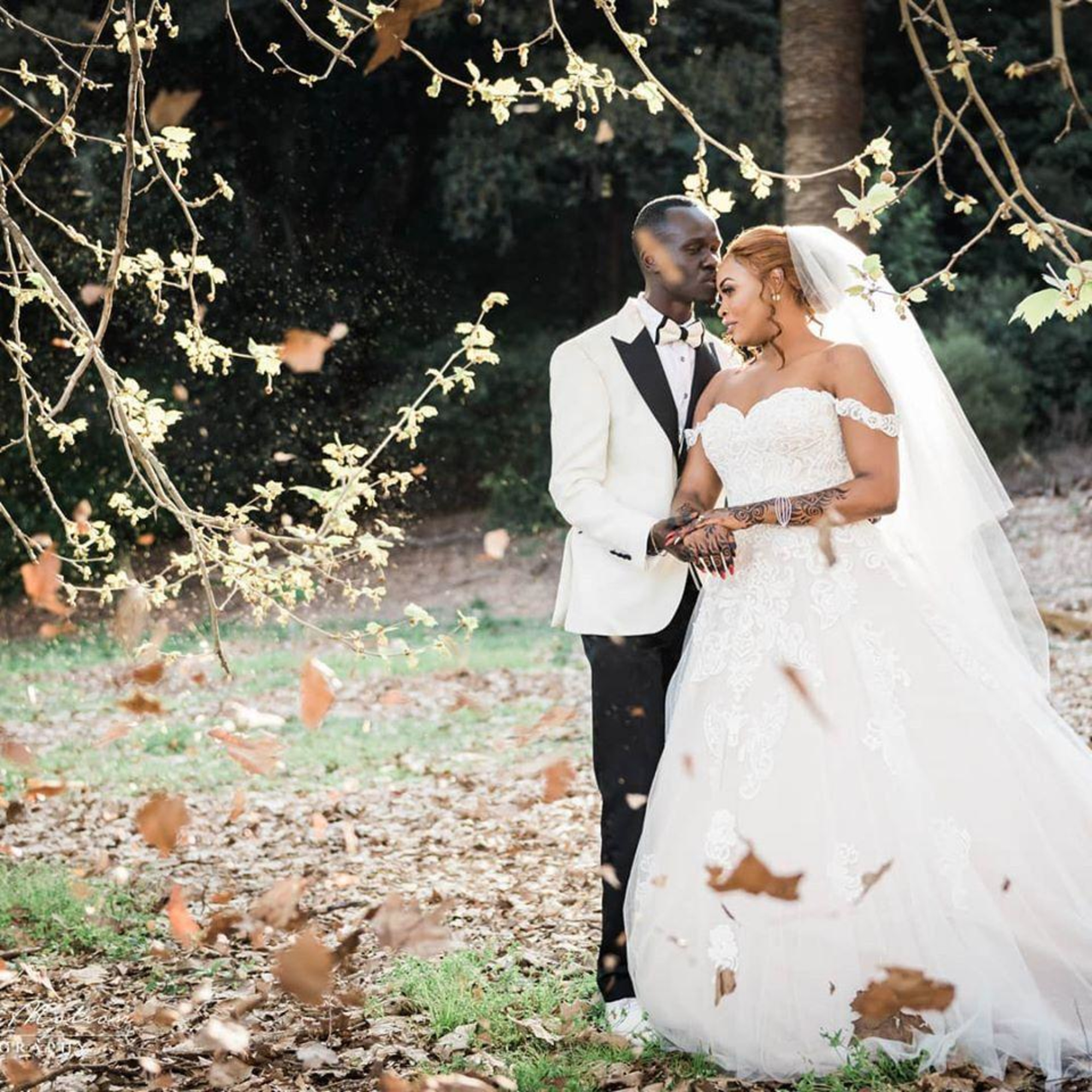 Bride and groom embrace in a leafy outdoor setting with falling autumn leaves around them.