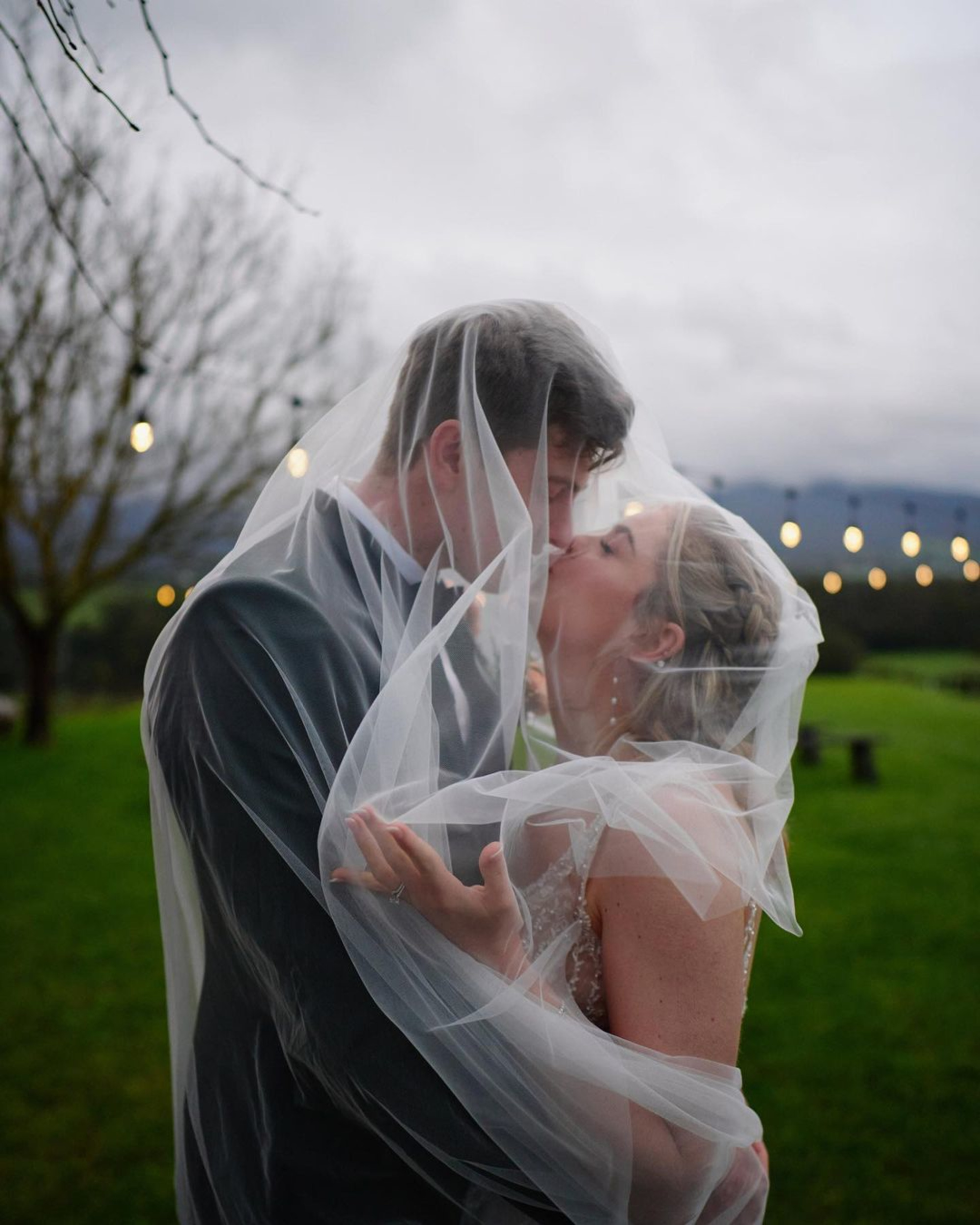 Bride and groom share a kiss under the veil outdoors at dusk with string lights in the background.