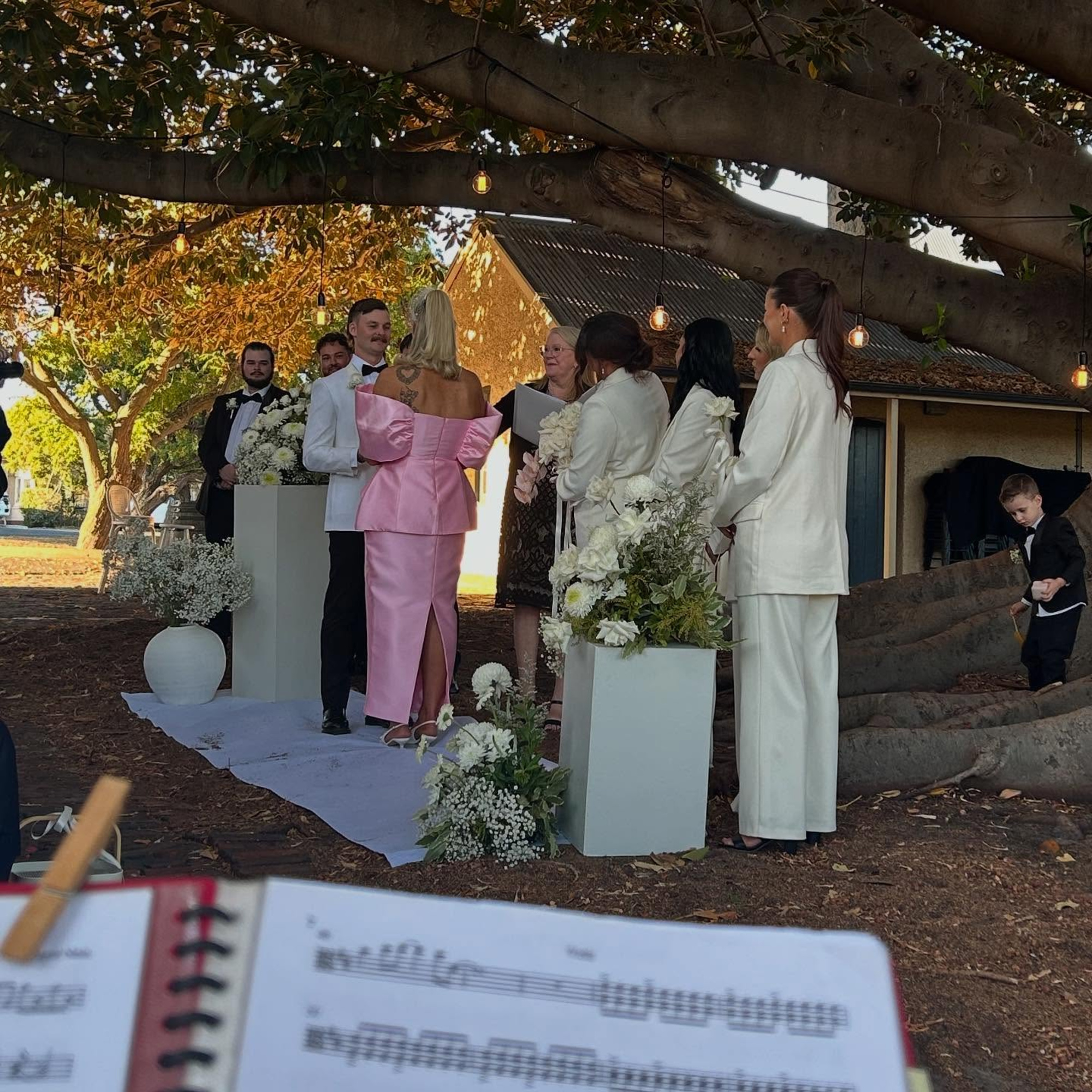 Outdoor wedding ceremony under a large tree with the couple exchanging vows surrounded by the bridal party and white florals.