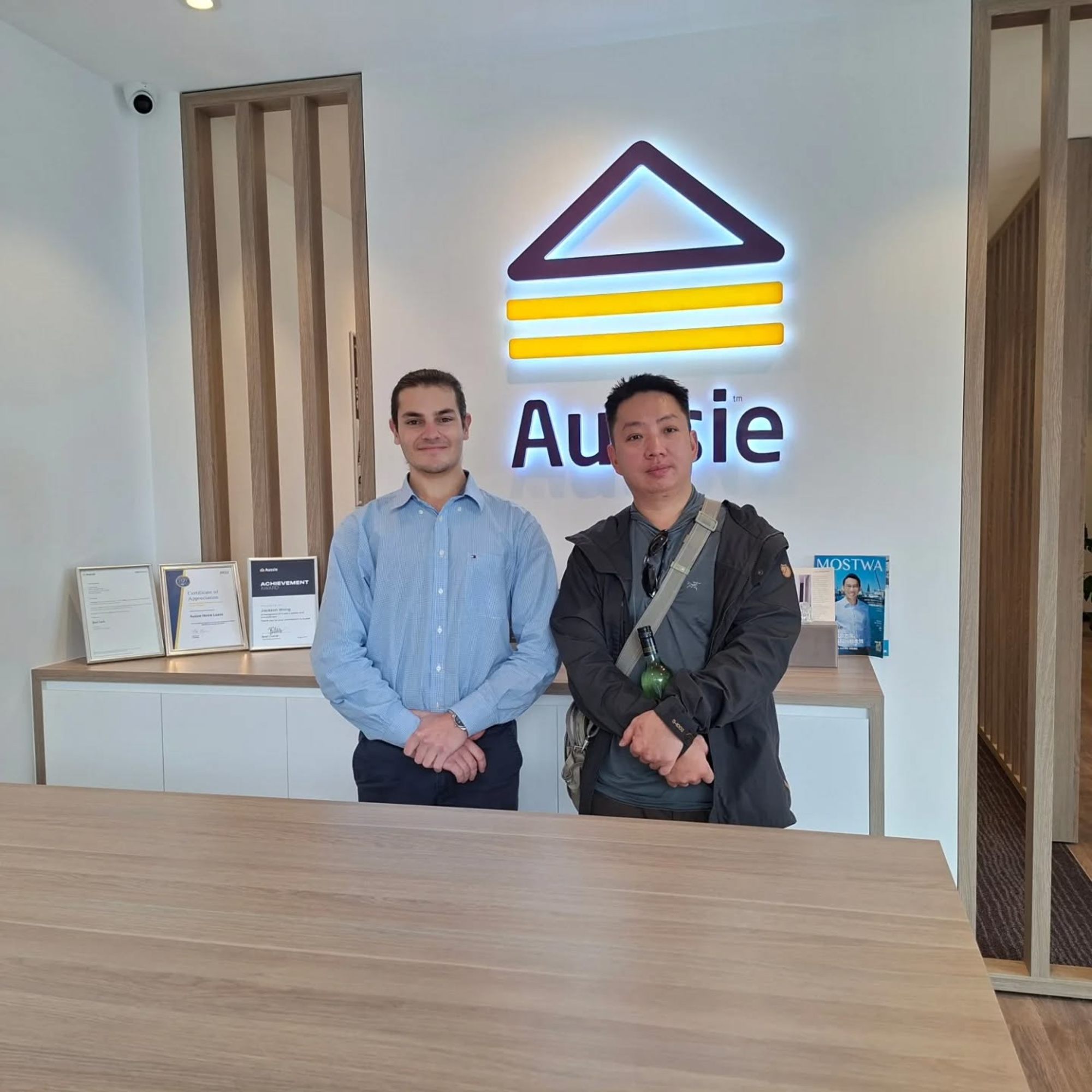 Two men stand in a modern office reception area in front of a lit wall logo.