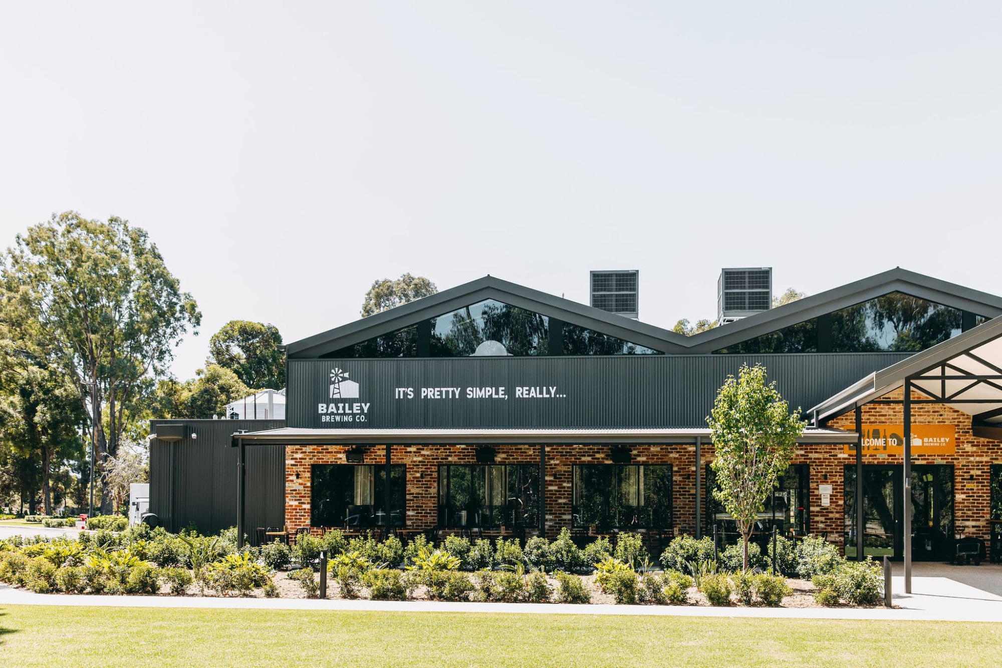 Modern brewery building with brick and metal exterior surrounded by landscaped gardens on a sunny day.