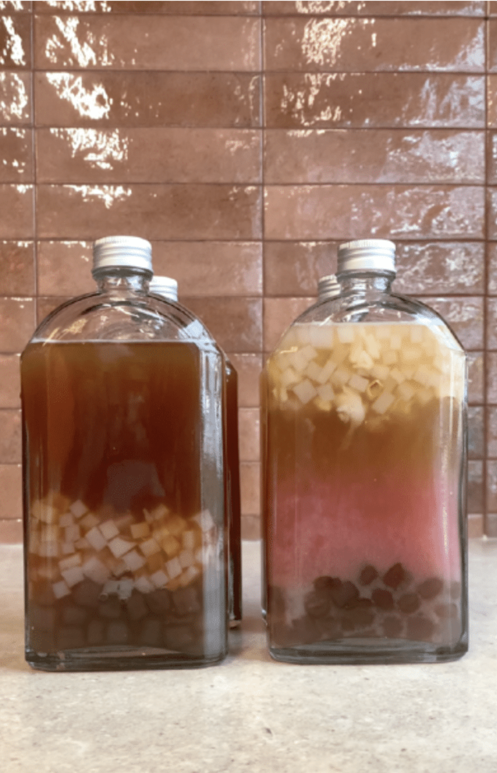 Bottled bubble tea drinks with fruit cubes and pearls arranged on a countertop against a tiled wall.