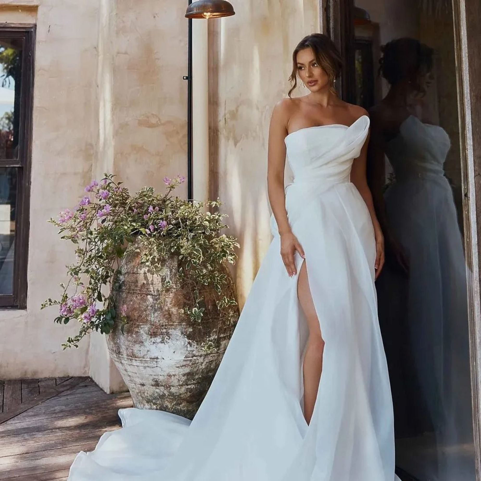 Bride in a strapless white gown with a thigh-high slit posing beside a rustic urn with flowers.