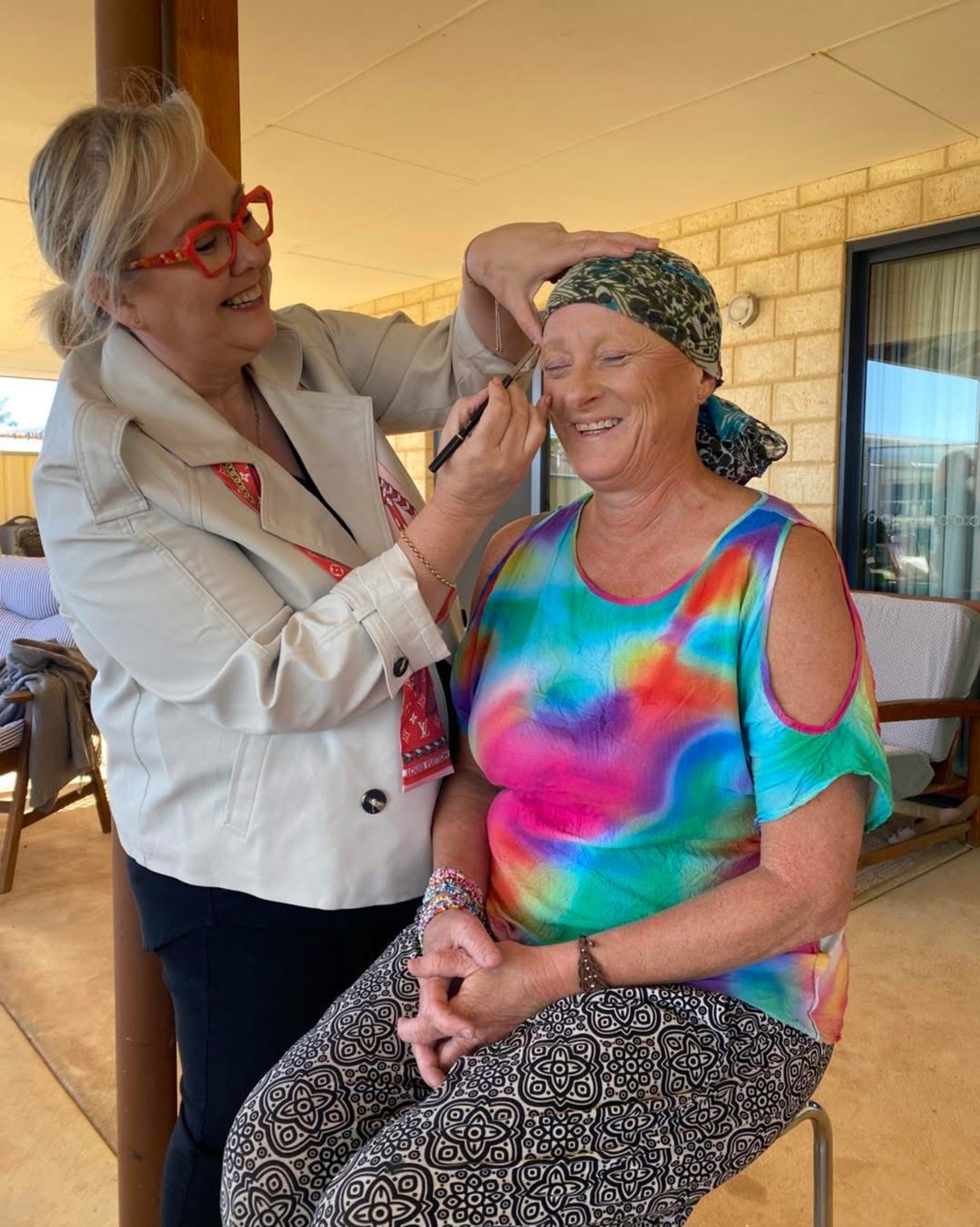 Makeup artist smiling while applying eye makeup to a seated woman in colorful clothing on a patio.