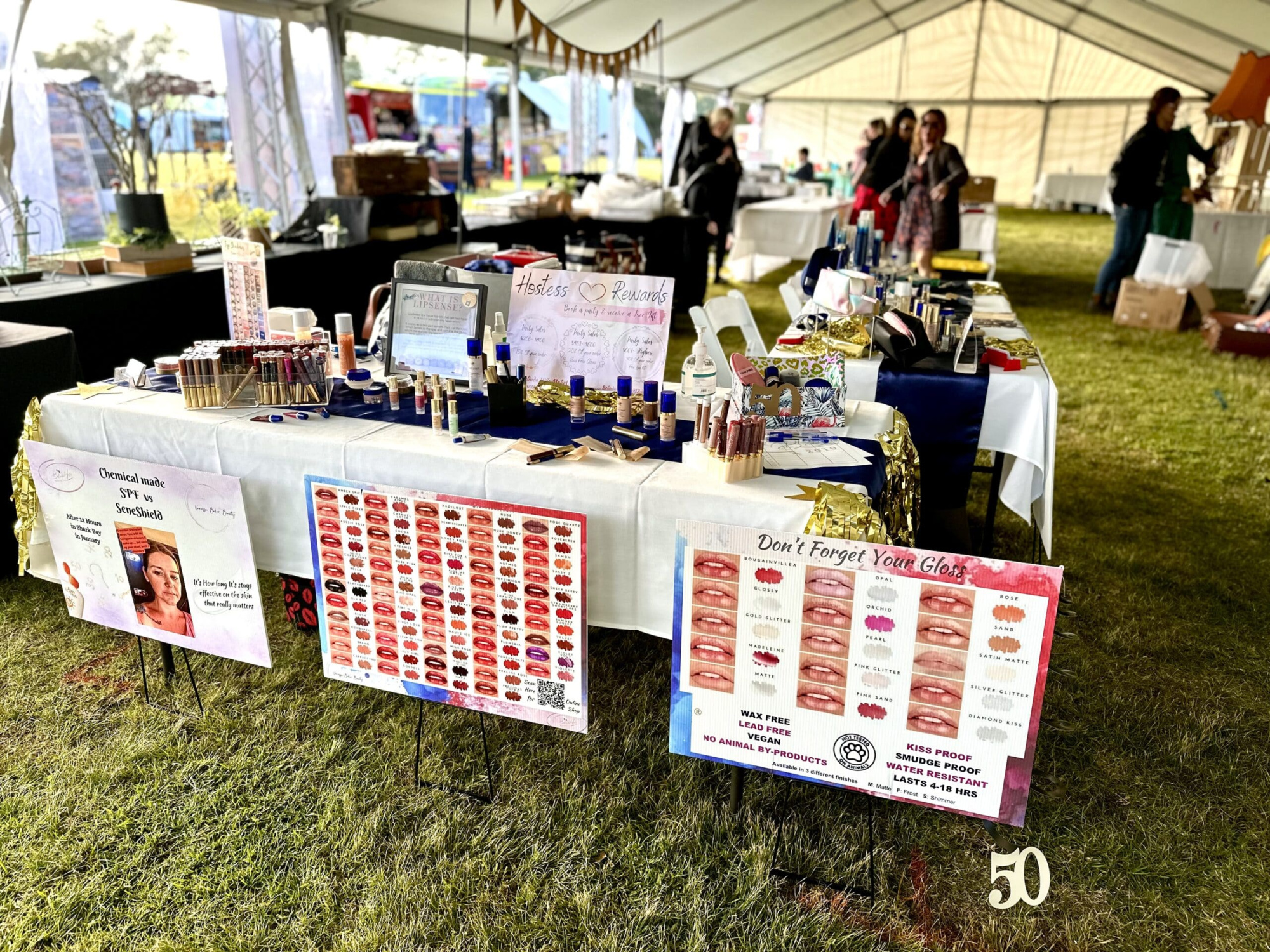 Outdoor bridal fair vendor booth displaying a variety of wedding makeup and lip gloss products on tables.