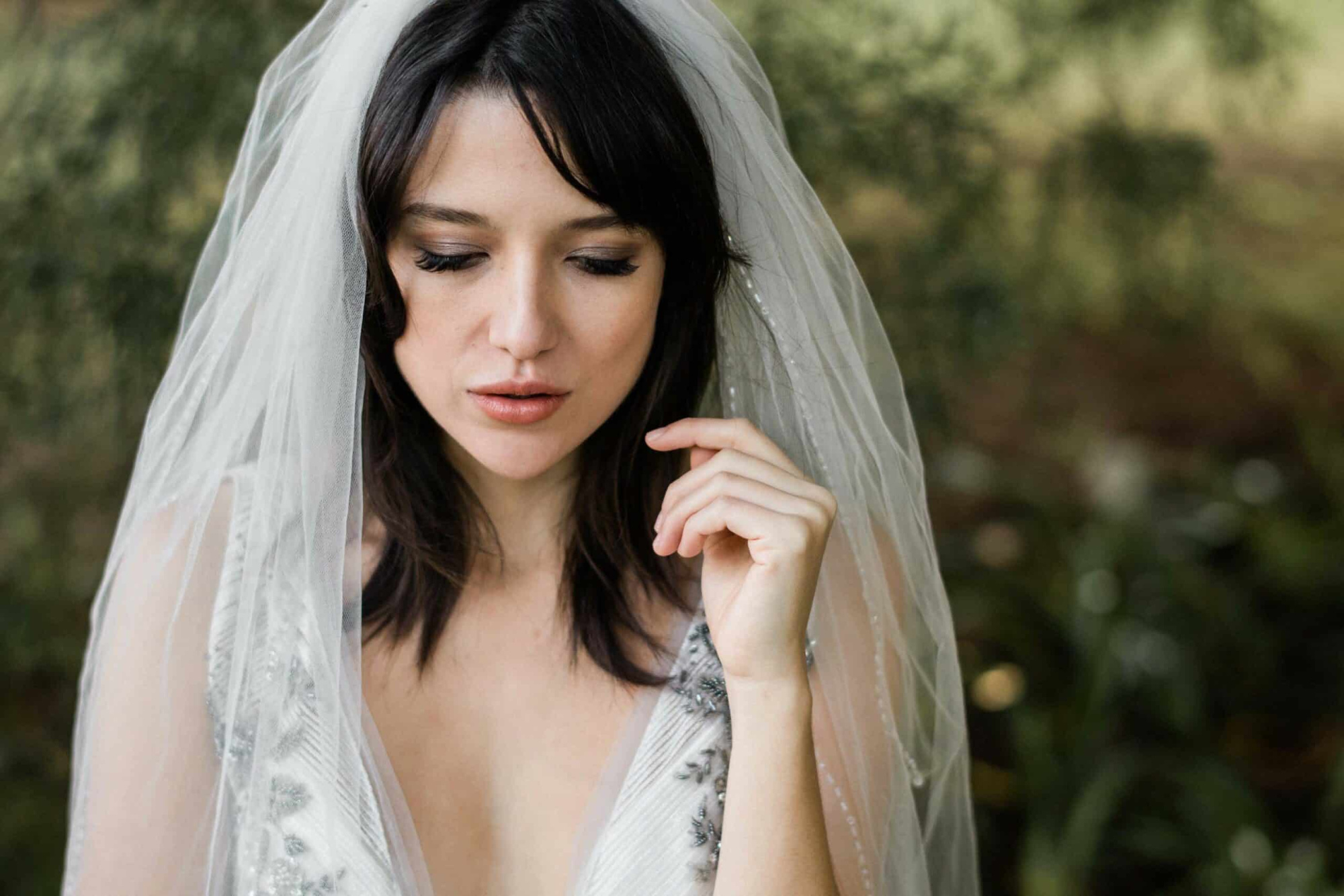 Close-up outdoor portrait of a bride in a beaded gown and veil softly touching her face.