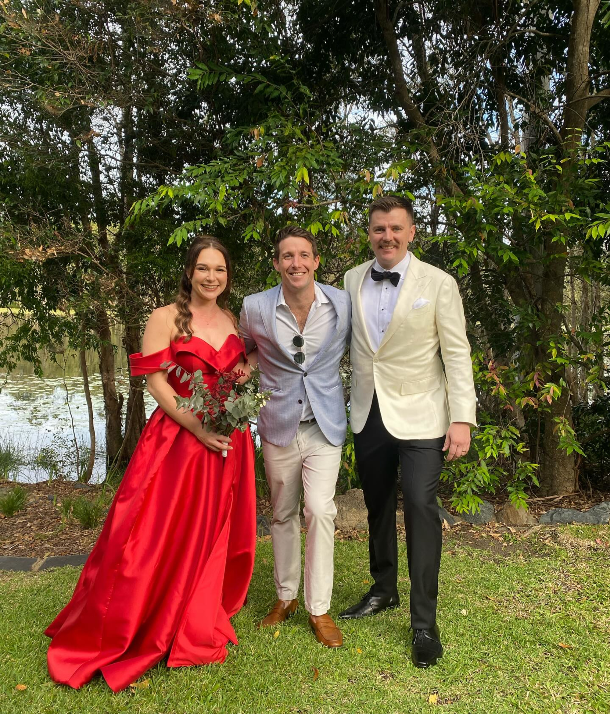 Three well-dressed wedding guests pose together outdoors in a lush garden setting.