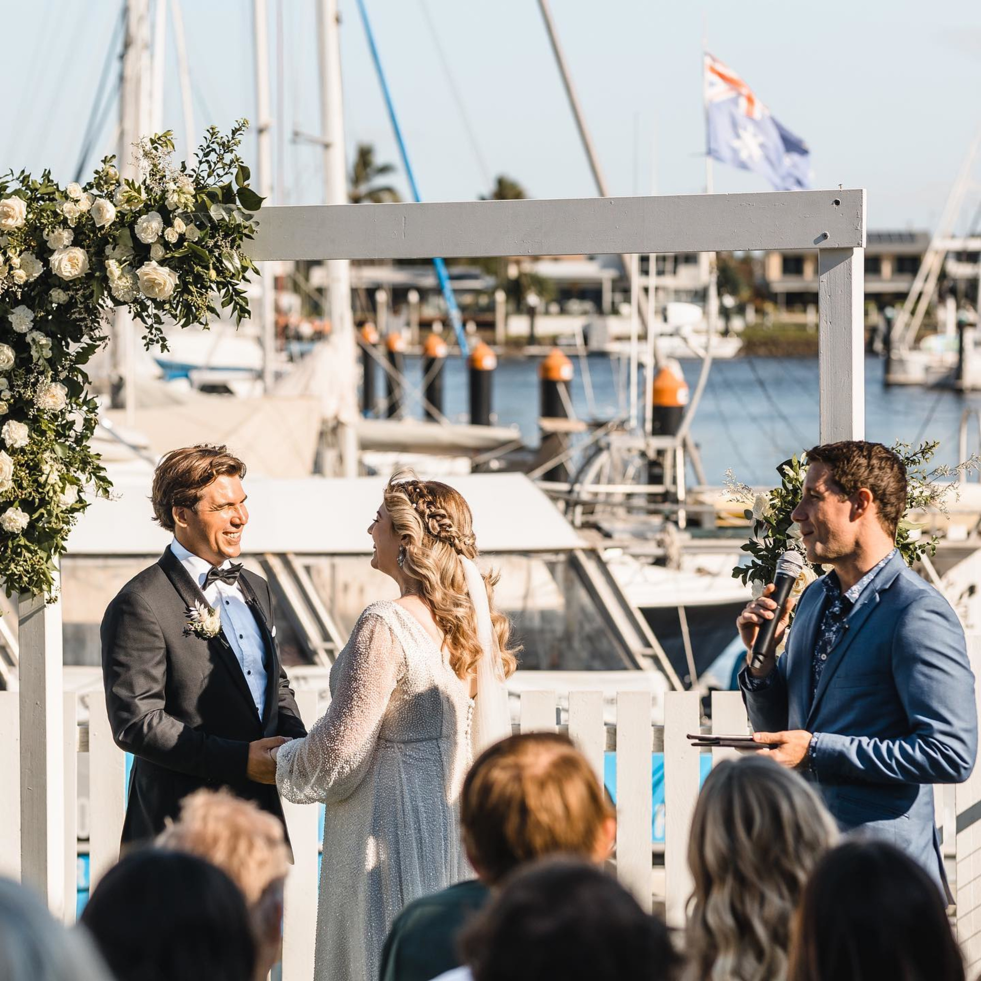 Outdoor marina wedding ceremony with couple holding hands under a floral arch as the celebrant speaks.