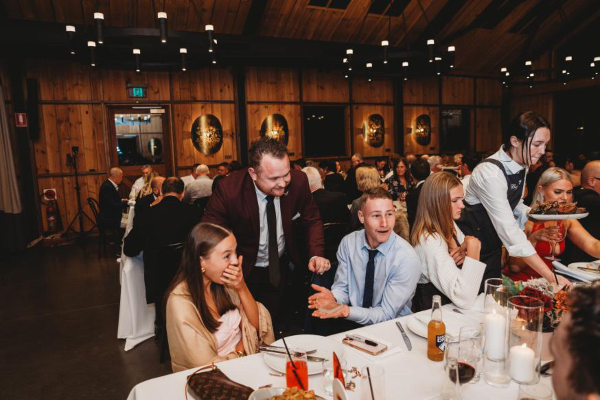 Guests enjoy dinner and entertainment at a lively indoor wedding reception in a warm wooden venue.