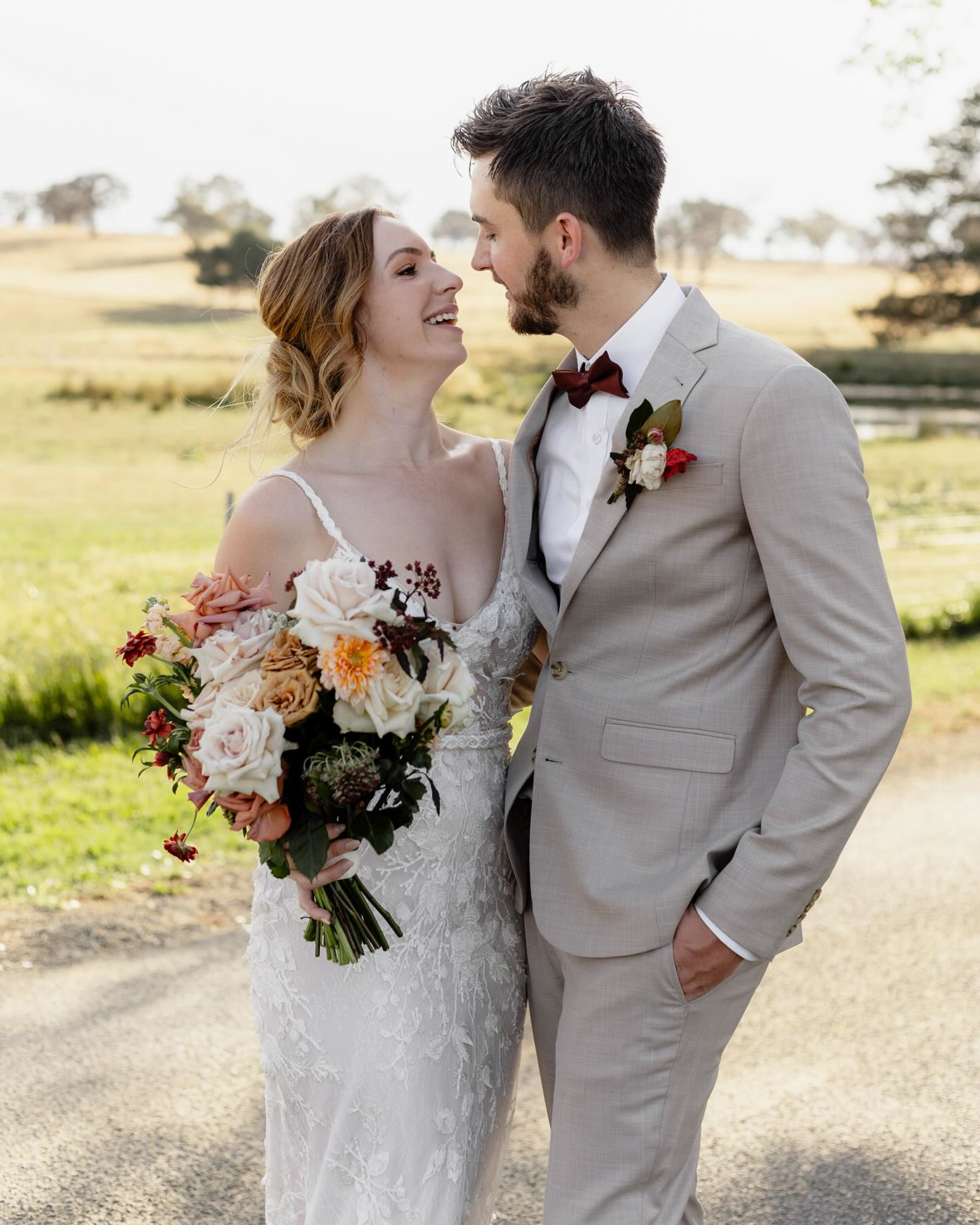 Bride and groom smiling at each other outdoors, holding a large bouquet of roses and seasonal flowers.