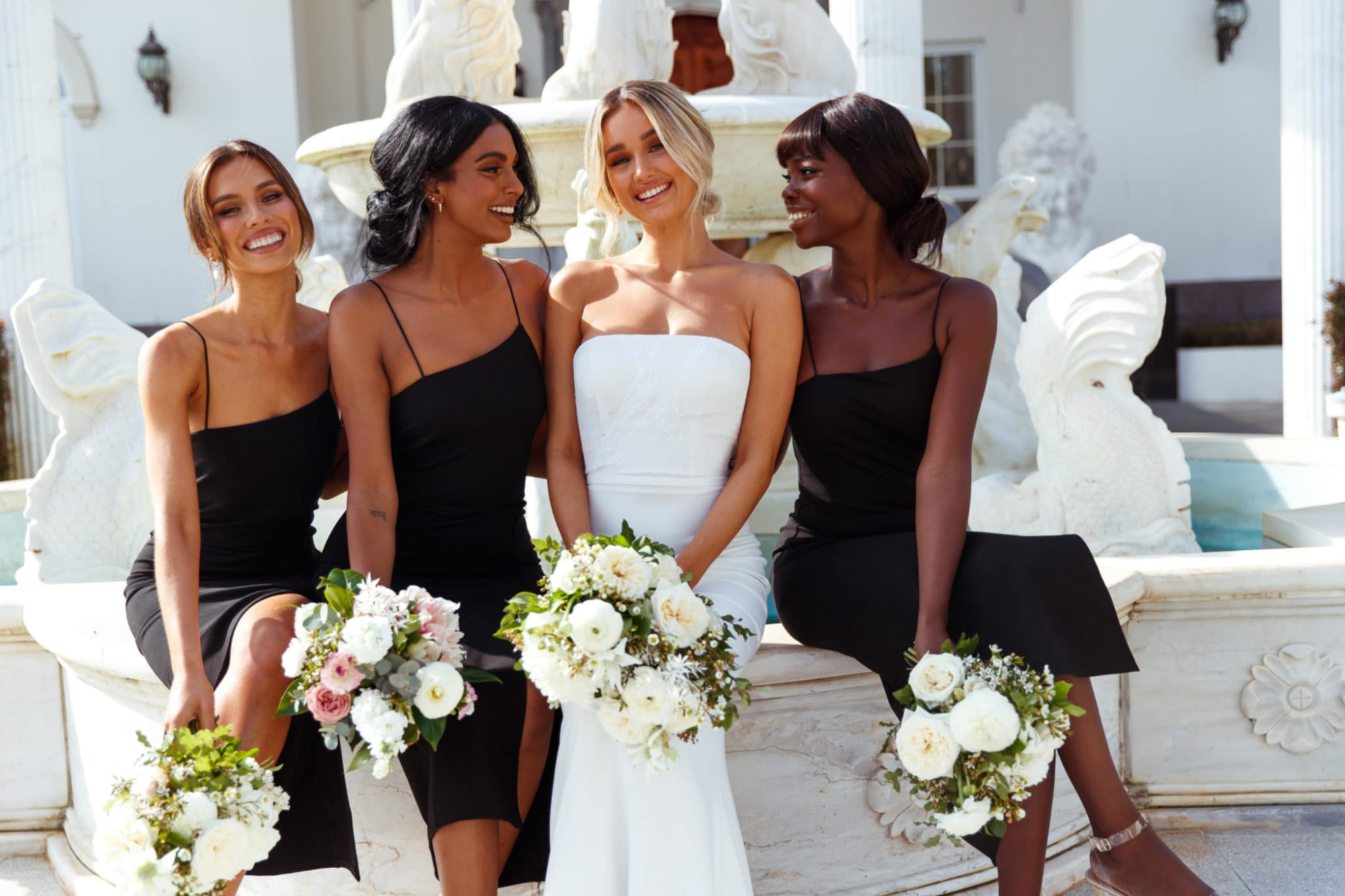 Bride in a white gown sits with three bridesmaids in black dresses holding white bouquets in front of a fountain.