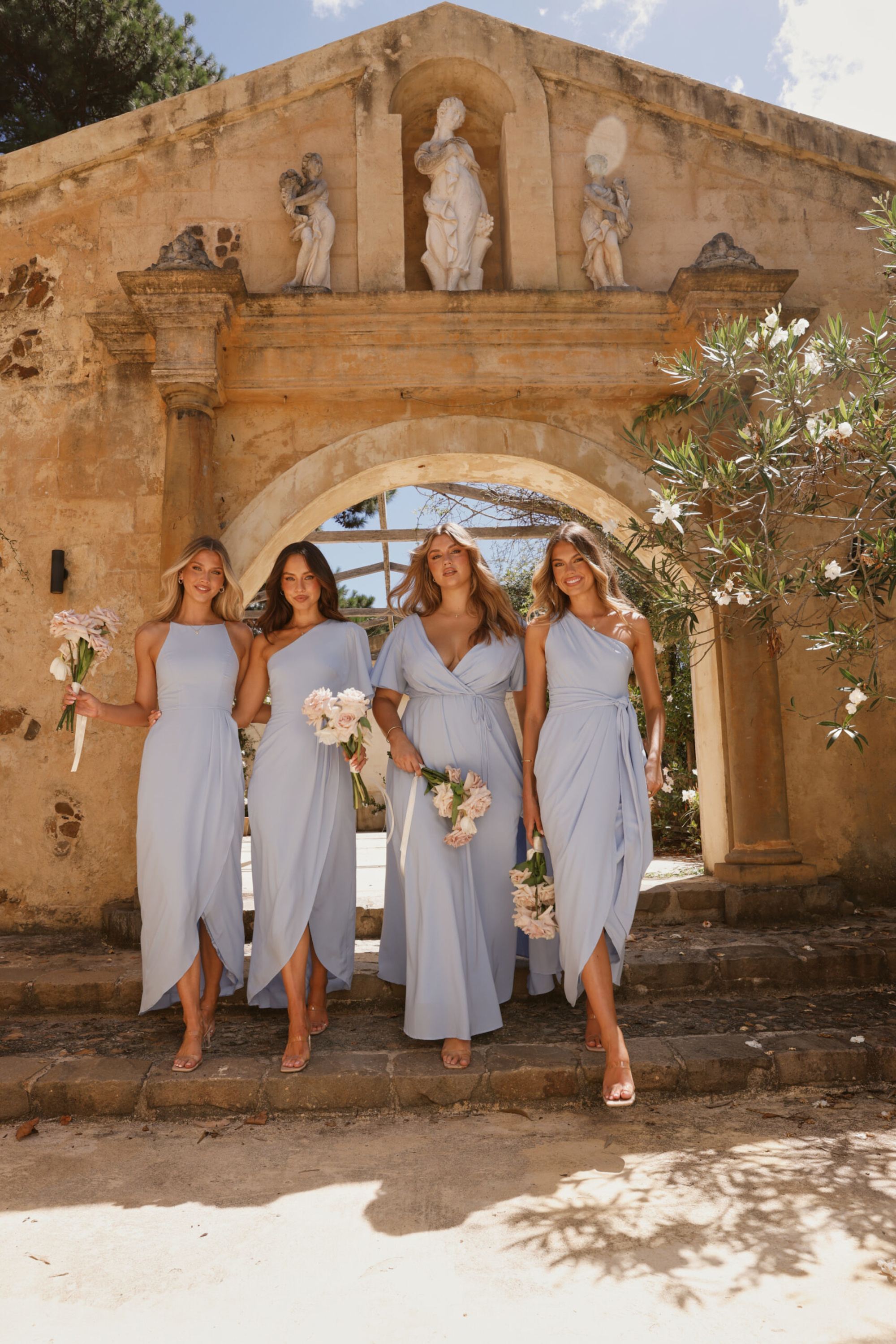 Four bridesmaids in light blue dresses holding bouquets walk under a rustic stone archway at an outdoor wedding venue.