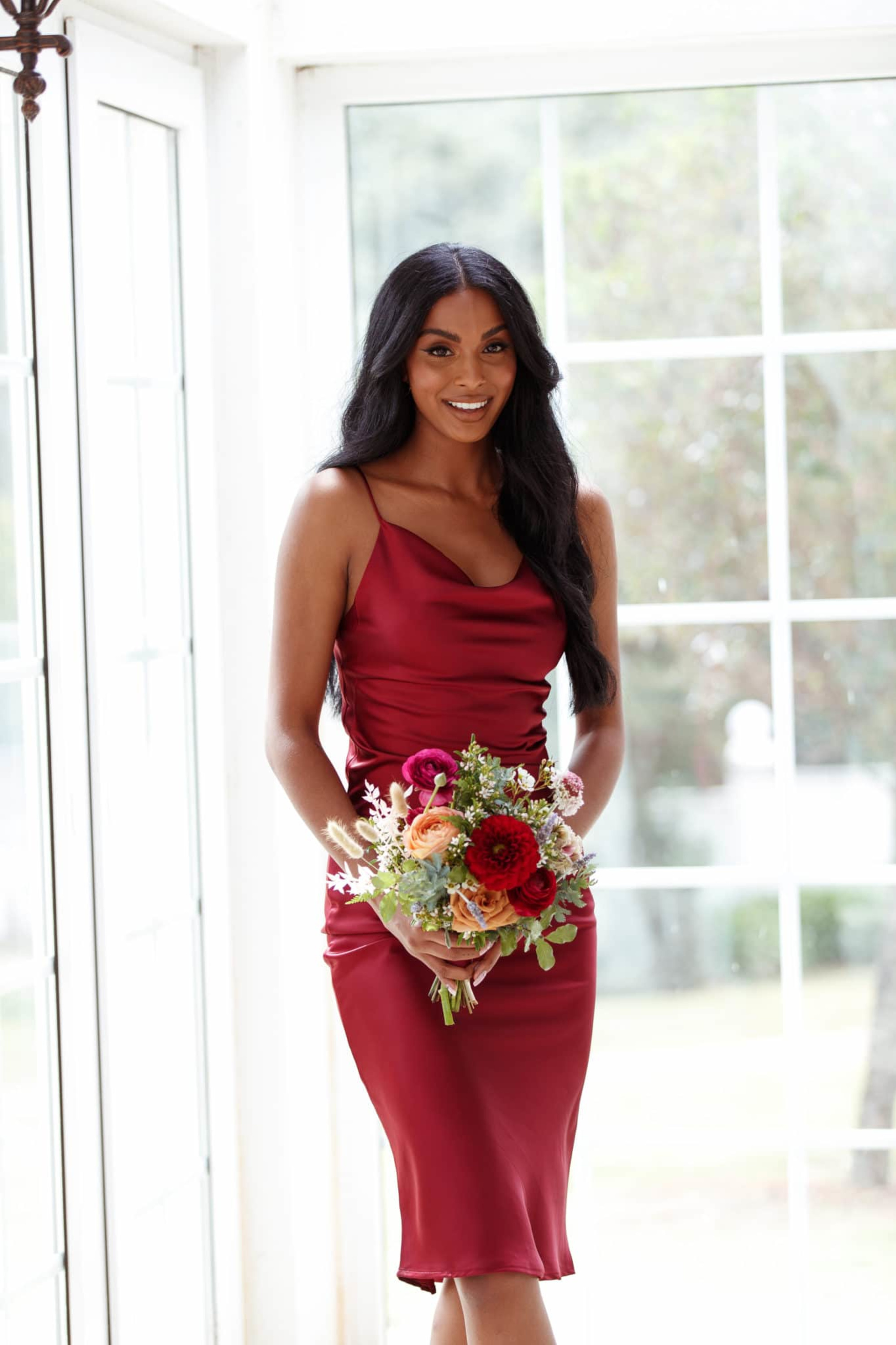 Bridesmaid in a satin burgundy dress holding a colorful bouquet in a bright indoor space.