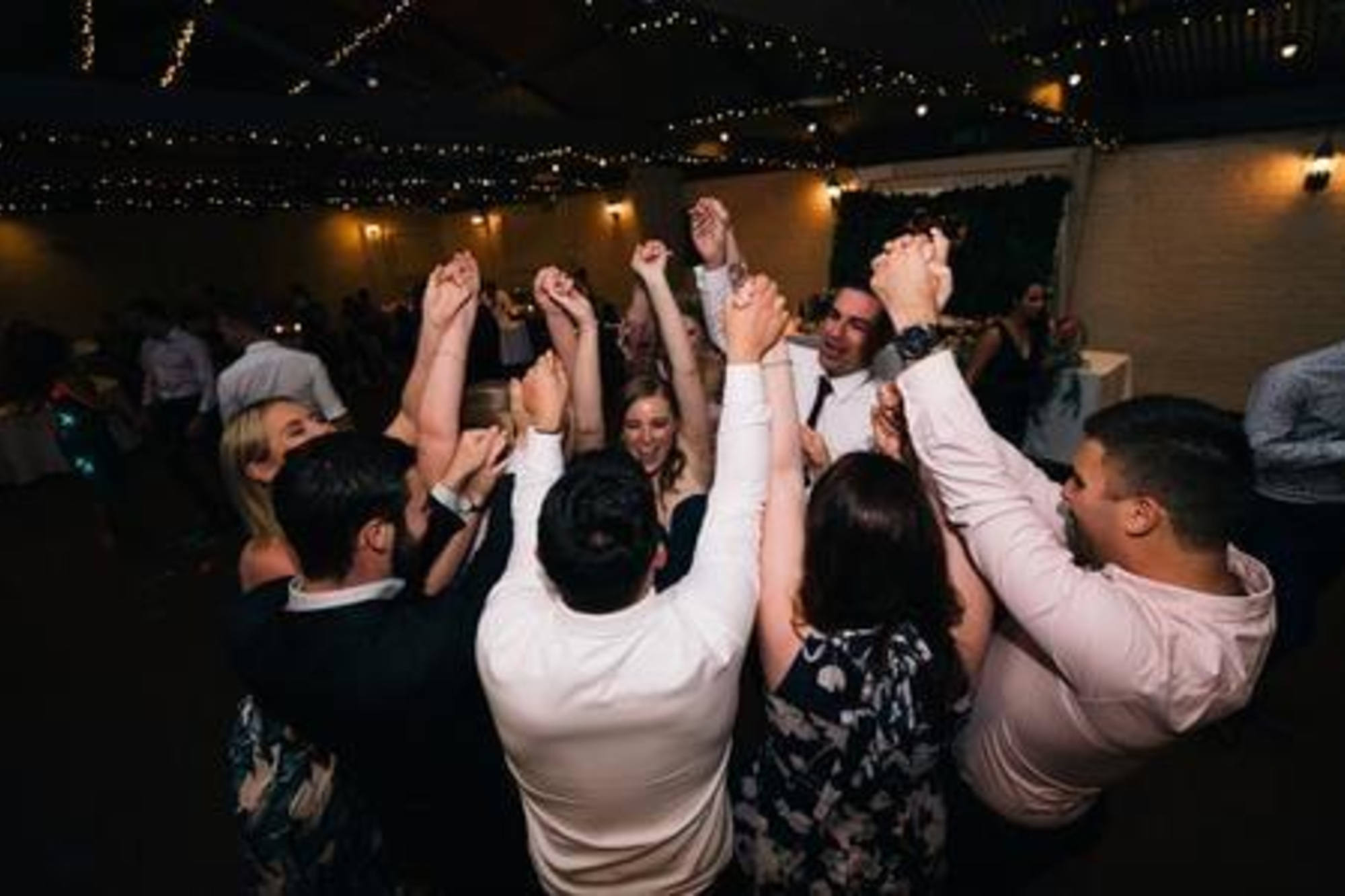 Wedding guests form a circle with raised hands dancing under string lights at an indoor reception.
