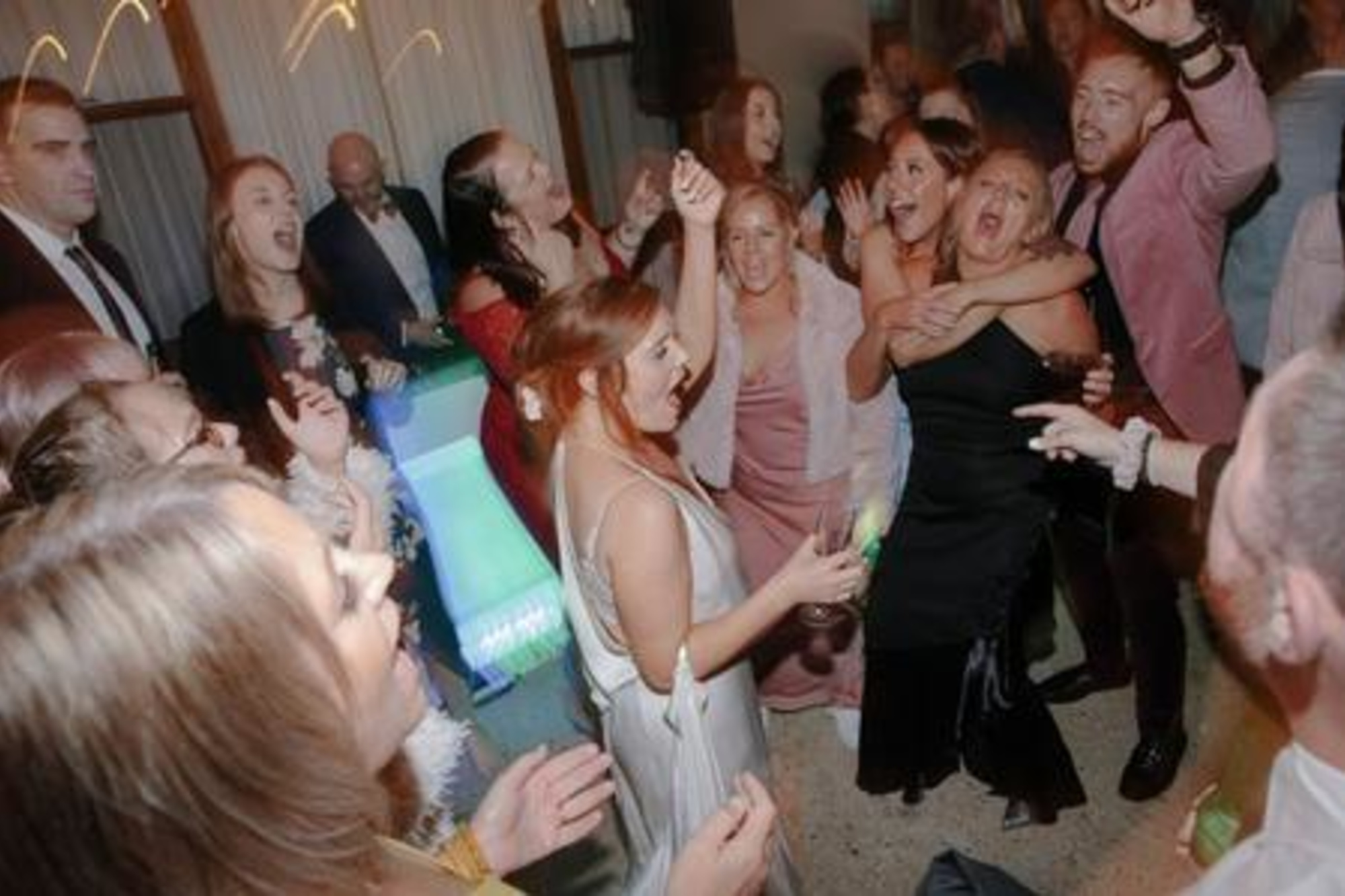 Wedding guests crowd the dance floor, singing and dancing together during a lively reception.